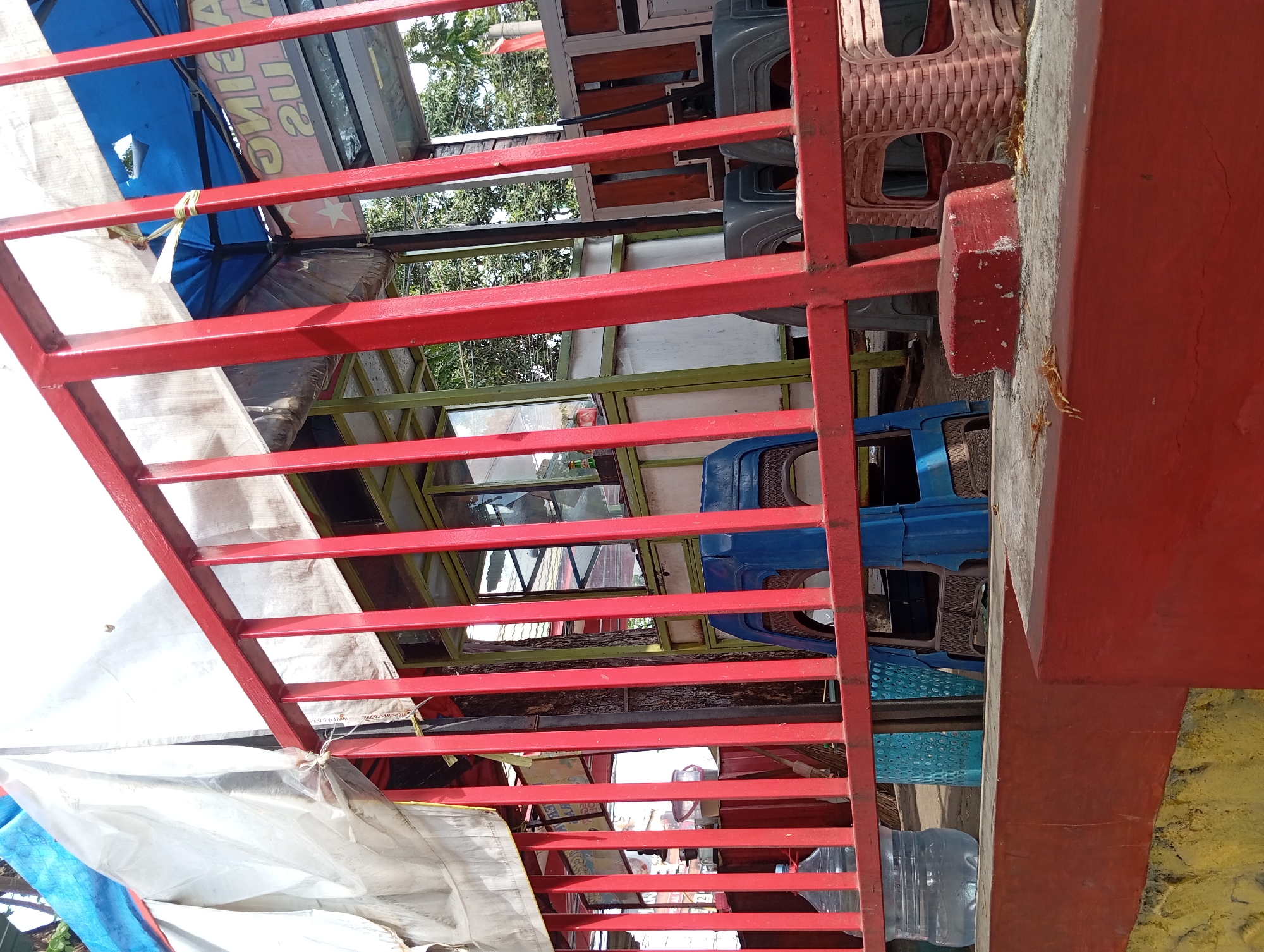 The image shows a roadside stall in Pandaan, Indonesia. A red metal fence runs across the foreground, obscuring part of the view of the stall. Behind the fence, there are several plastic chairs, a large water bottle, and a broom leaning against a stall. The stall itself appears to be constructed of wood and glass, with a blue tarp overhead. A sign with Indonesian text is visible on the stall. Trees and buildings can be seen in the background, suggesting an urban or semi-urban environment. The lighting indicates it is daytime, likely sunny given the bright white of the tarp. There are no people visible in the image.