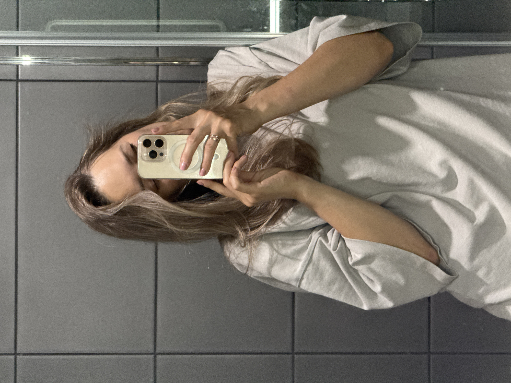 A woman is taking a mirror selfie in a bathroom. She has long, light brown hair and is wearing a light gray oversized t-shirt. She is holding a smartphone in her right hand, which is obscuring part of her face. A ring is visible on her right ring finger. The background consists of gray tiled walls and a glass shower door. The scene is brightly lit. The image is likely taken indoors in a private residence or hotel. The provided crops offer close-ups of the woman's hair, hands, phone, and clothing, as well as the surrounding tiled wall. There is no information in the image to indicate the location as Chaikovsky, Russia.
