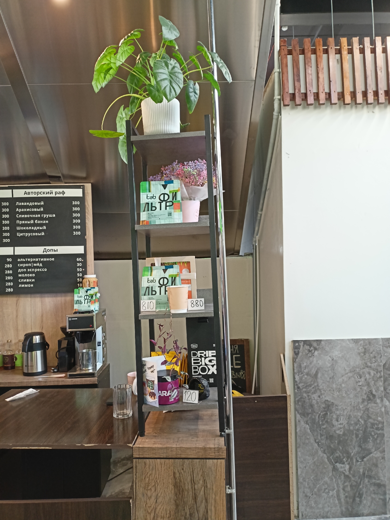 Here is a description of the image:
**Scene:** The photograph appears to be taken inside a cafe or coffee shop. The location is likely in Novosibirsk, Russia, as indicated by the Cyrillic script on the menu and product packaging.
**Objects:**
* A chalkboard menu with Cyrillic writing, listing different coffee drinks and add-ons with prices.
* A multi-tiered shelf unit made of a dark metal frame and shelves, holding various items:
* A green leafy plant in a white pot at the top.
* Branded packaging and merchandise with Cyrillic script.
* Flowers.
* Coffee cups.
* A small jar.
* Coffee-making equipment (a coffee maker and a grinder) on a countertop.
* A wooden countertop.
* A glass.
**Environment:** The cafe appears modern with stainless steel and wooden surfaces.