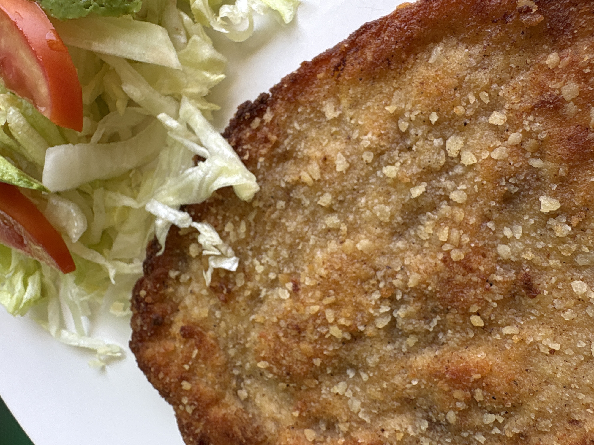 This close-up image showcases a dish consisting of a large, golden-brown, breaded cutlet served alongside a simple side salad on a white plate.
The main subject is the crispy, fried cutlet, which takes up a significant portion of the frame. Its surface is textured with visible breadcrumbs or a similar coating, some of which appear to be lightly toasted or seasoned flakes, possibly grated cheese, adding to its appealing golden and reddish-brown color. The edges of the cutlet are well-defined and slightly darker, indicating a good crisp.
To the left and top-left of the cutlet, a fresh salad is visible. It primarily consists of shredded light green lettuce. Nestled among the lettuce are a couple of bright red tomato slices and what appears to be a slice of green avocado at the very top. The lettuce and tomatoes have a fresh, slightly glistening appearance.
The food is presented on a clean, white ceramic plate, which occupies the lower left corner and extends behind the food items. The background beyond the plate is out of focus, showing a dark green hue, possibly a placemat or table surface.
The image captures the dish from a slightly elevated, close-up perspective, emphasizing the textures and colors of the food. The scene suggests a meal ready to be eaten, likely indoors in Mexico City, Mexico, given the context. There are no people visible, nor any discernible text. The focus is entirely on the culinary presentation.