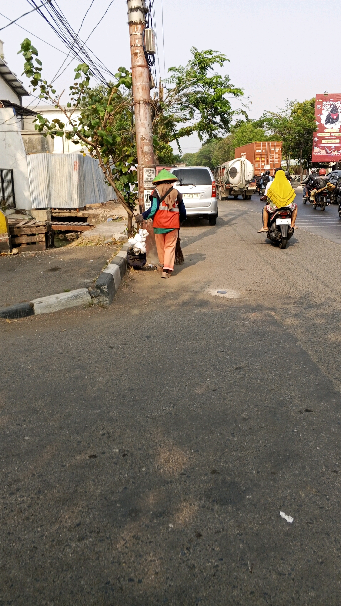 The image shows a street scene in Semarang, Indonesia. In the foreground, a street cleaner wearing an orange vest and a green conical hat sweeps refuse with a broom. They are standing next to a metal bin and a small tree. A silver van is parked behind the street cleaner. In the background, several motorcycles are visible, along with a white truck with a water tank and a large orange shipping container on a truck. A billboard advertising "Universitas" with a bird logo is on the right side of the image. The scene appears to be a busy roadside with a mix of vehicles and pedestrians.