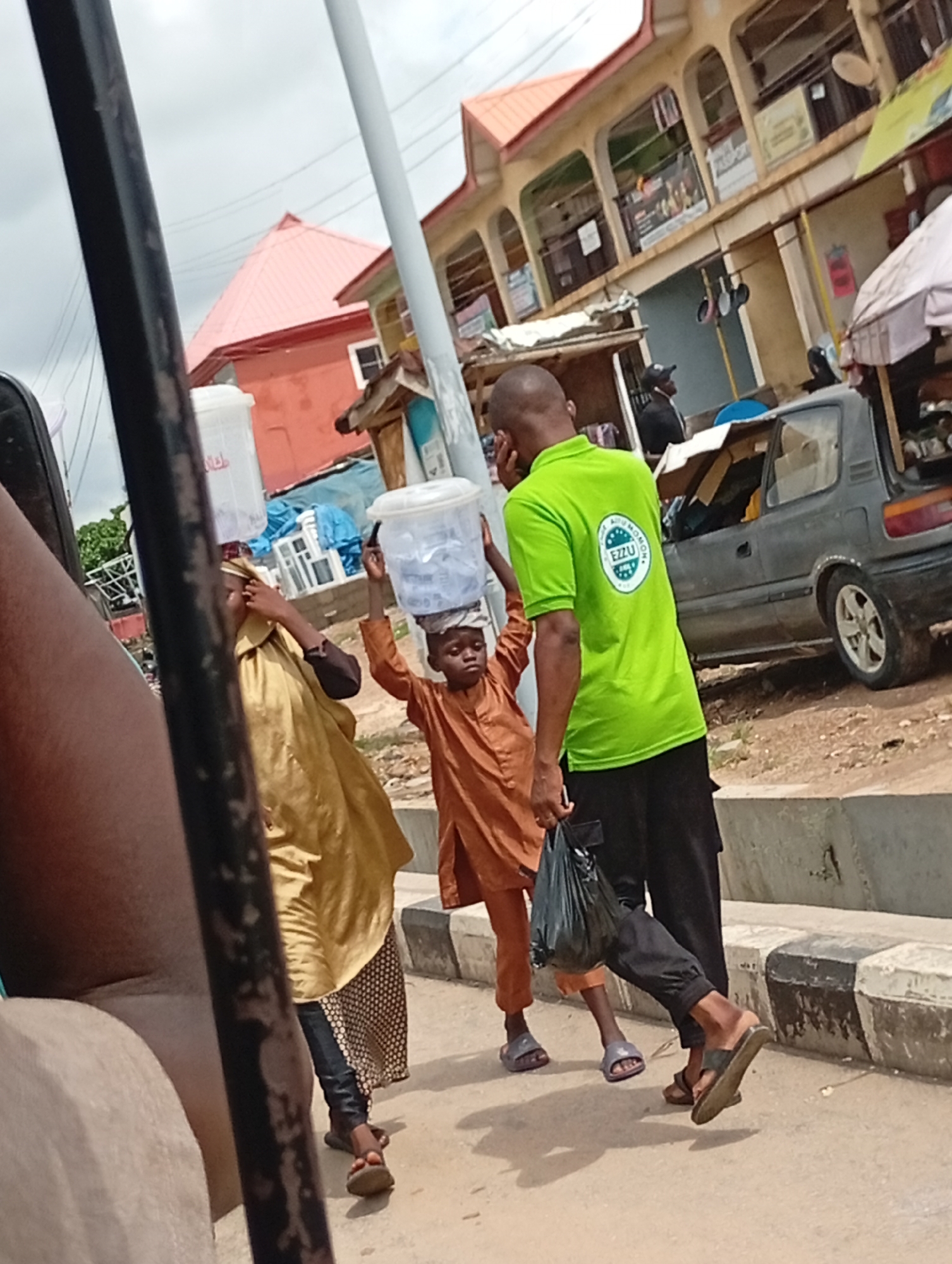 The image depicts a street scene in Lokoja, Nigeria. A woman wearing a yellow gown with black leggings and a brown head covering is carrying a white bucket on her head. A young boy wearing a brown tunic and pants is also carrying a white bucket on his head. A man is walking beside the boy, wearing a green shirt with a logo on the back and black pants. He is carrying a black plastic bag. They are walking on a concrete sidewalk next to a street with a gray car parked on the side. In the background is a two-story building with a red roof and a commercial area. A utility pole is located near the people.