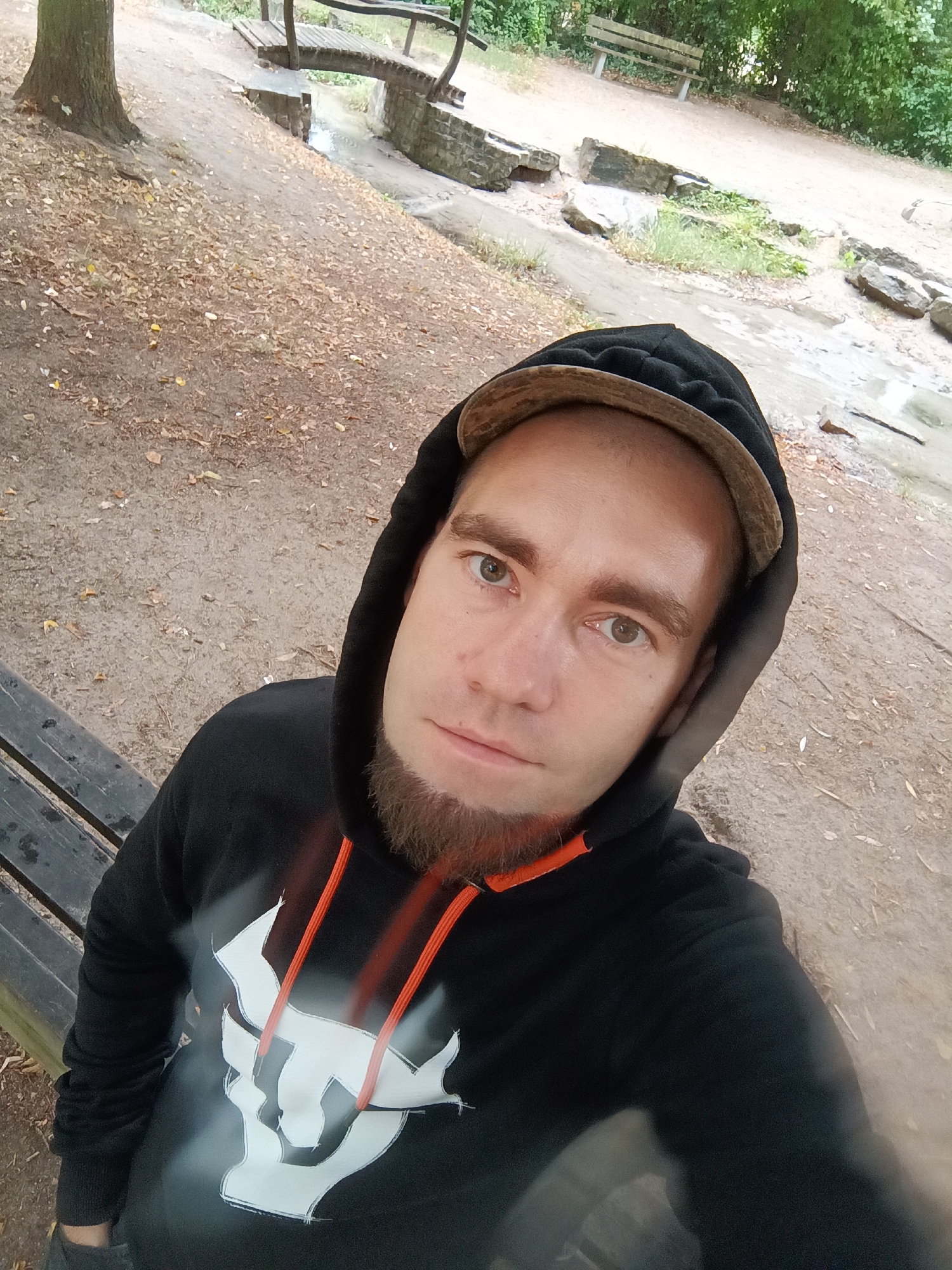 A man wearing a black hooded sweatshirt with orange drawstrings and a beige baseball cap is pictured outdoors. He has brown hair, a light brown beard, and light brown eyes. He is looking directly at the camera. The sweatshirt has a white graphic design on the chest. He appears to be sitting on a wooden bench.
The background shows a park-like setting with a dirt path, fallen leaves, trees, a small wooden bridge crossing a narrow stream, large rocks, and a wooden bench in the distance. The scene suggests an autumn or early spring environment. The presence of a stream and surrounding vegetation indicates a natural or semi-natural area, possibly within a city park in Schorndorf, Germany.