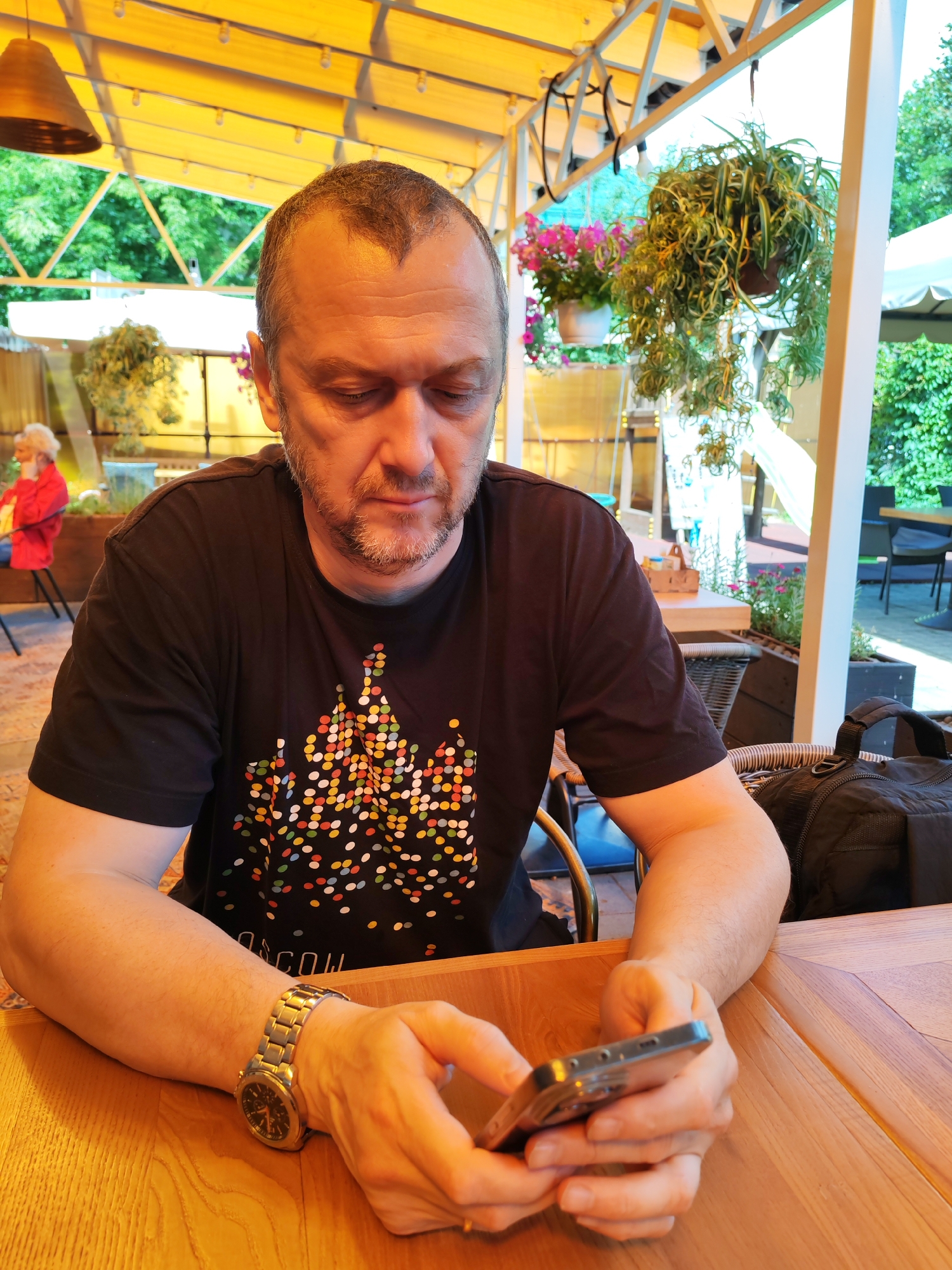 In this medium shot photograph, a man with short, receding brown hair and a goatee is seated at a wooden table, looking down at his smartphone. He is wearing a black t-shirt with a colorful, abstract pattern of dots. His left arm, adorned with a silver watch, rests on the table as his hands cradle the phone. To his right, a black duffel bag is placed on a woven chair. The background suggests an outdoor cafe setting, with a yellow awning overhead, hanging plants, and glimpses of other patrons and tables. The man appears to be in Bol'shie Mkhi, Russia, an area known for its natural beauty and outdoor recreational activities.