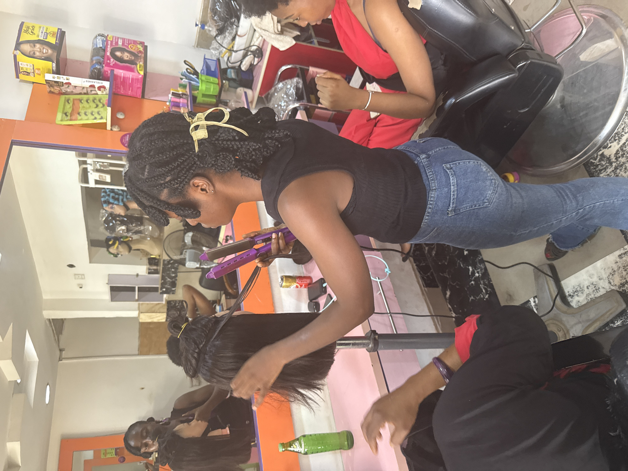 A woman in blue jeans and a black top is straightening black hair with a purple straightening iron at a hair salon in Abuja, Nigeria. Another woman in a red top is sitting in a black salon chair next to her. There are various hair products, boxes, and accessories visible on shelves and counters. A large mirror reflects other people in the salon.