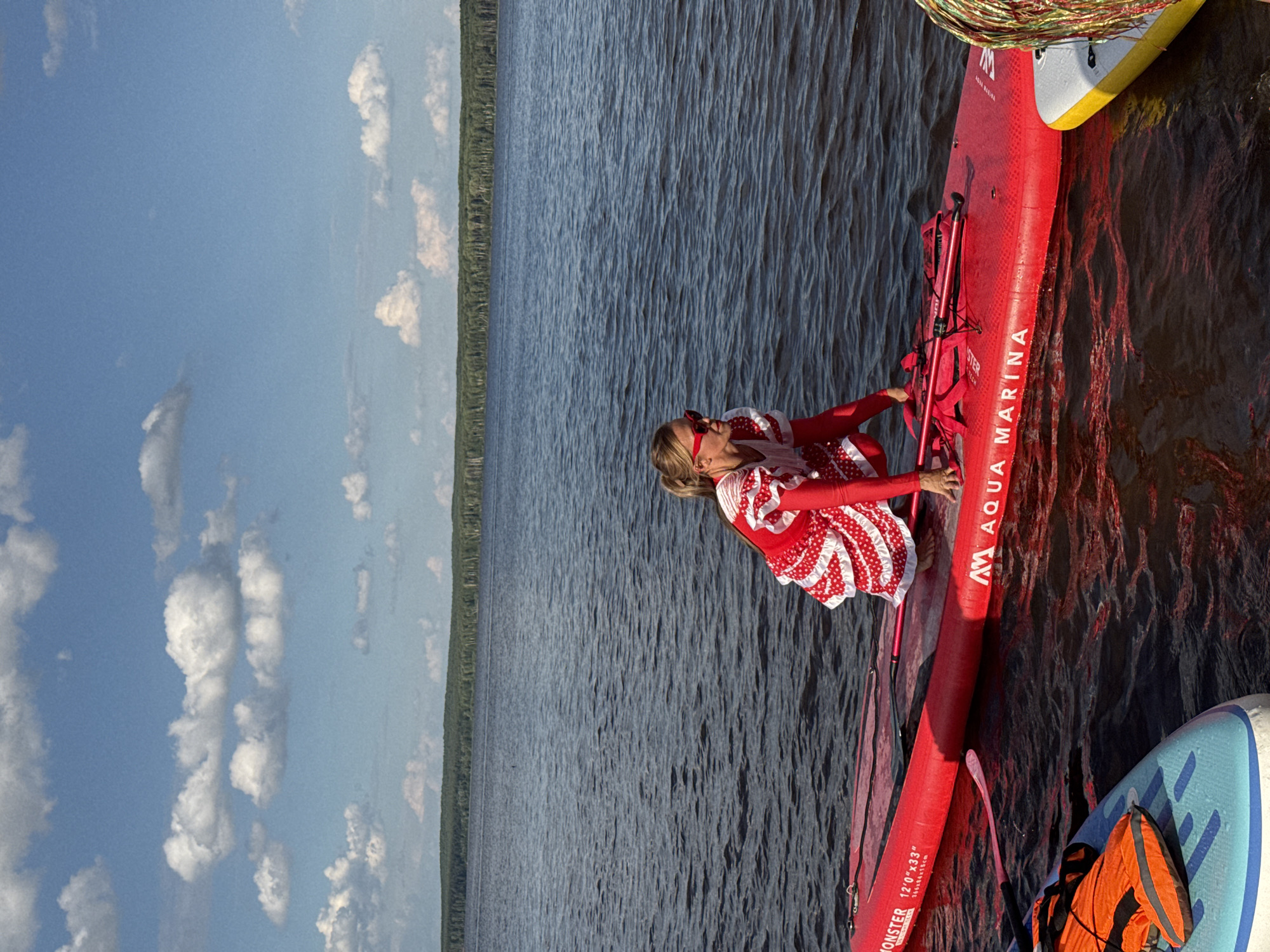 A woman wearing a red and white polka dot dress and red sunglasses squats on a red inflatable stand-up paddleboard in a dark body of water under a blue sky with scattered white clouds. The paddleboard has "AQUA MARINA" printed on its side. Another paddleboard, light blue with an orange life vest on it, is to the left of the red paddleboard. A third paddleboard, yellow and white, is to the right of the red one. The background shows a dense line of trees along a distant shore. The image is taken from a medium distance, showing the woman, the paddleboards, and the water. The location is the Kama River in Chaikovsky, Russia.