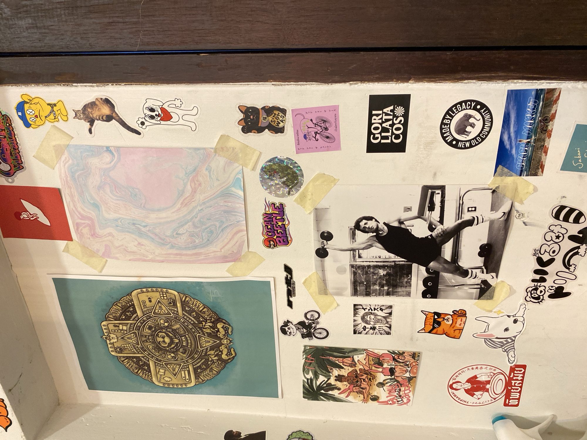This image shows a collage of stickers and images attached to a wall, likely in a restroom stall given the edge of a sink visible at the bottom. The stickers feature a variety of themes, including animals, brand logos, cartoons, and cultural imagery. The subjects include cats, dogs, a person lifting weights, and various animated figures. Specific stickers include: "GORI LLATA COS," "MADE BY LEGACY," "CHACHA MARKET," and "HIPSAMAI - THIPSAMAI," which is related to a restaurant in Bangkok, Thailand, known for Thai food, specifically "Pad Thai". A circular image features an ancient Aztec-style design. There is also a black-and-white photo of a person lifting weights in what appears to be a home gym setting. The stickers and images are adhered to the wall using tape.