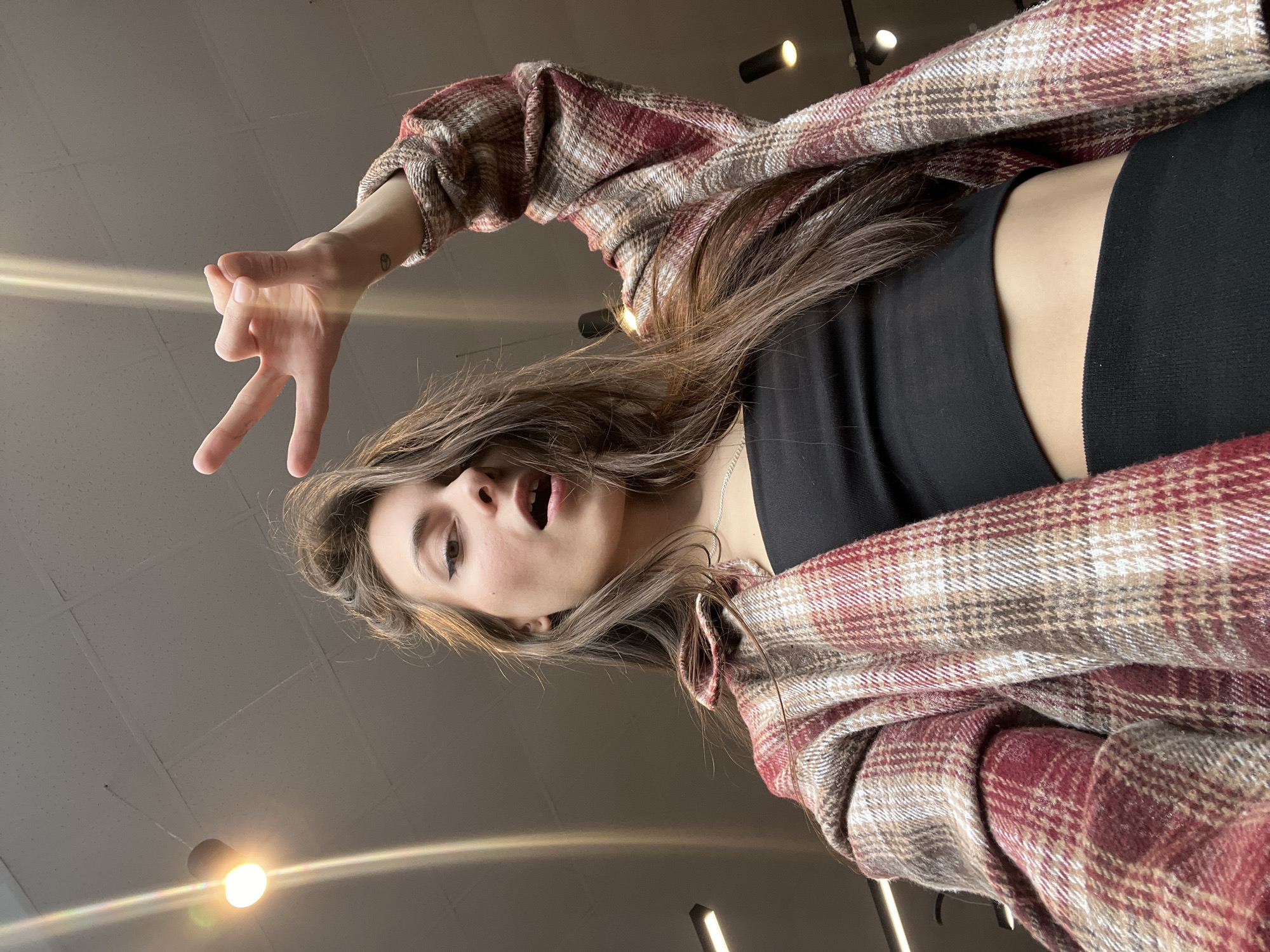 A young woman with brown hair and light skin is angled downward in the frame, wearing a black top and a red and brown plaid shirt. She is making a hand gesture, possibly a peace sign, with her left hand. Her mouth is open in an expression. Above her, several light fixtures are visible on the ceiling. The location appears to be indoors and the lighting suggests a modern or commercial space. There is no explicit evidence in the image to confirm its location in Krasnoyarsk, Russia.