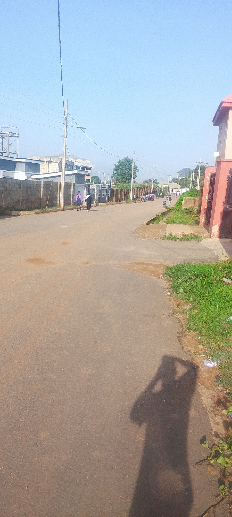 In the city of Jos, Nigeria, a paved road stretches into the distance under a clear blue sky. On the left side of the road, a brick wall borders several buildings, with a tall pole and power lines overhead. A few individuals are walking along the road, some appearing to be students in uniforms. On the right side, overgrown grass and a red-walled building are visible. The shadow of a person holding a phone or camera is cast on the road in the foreground.