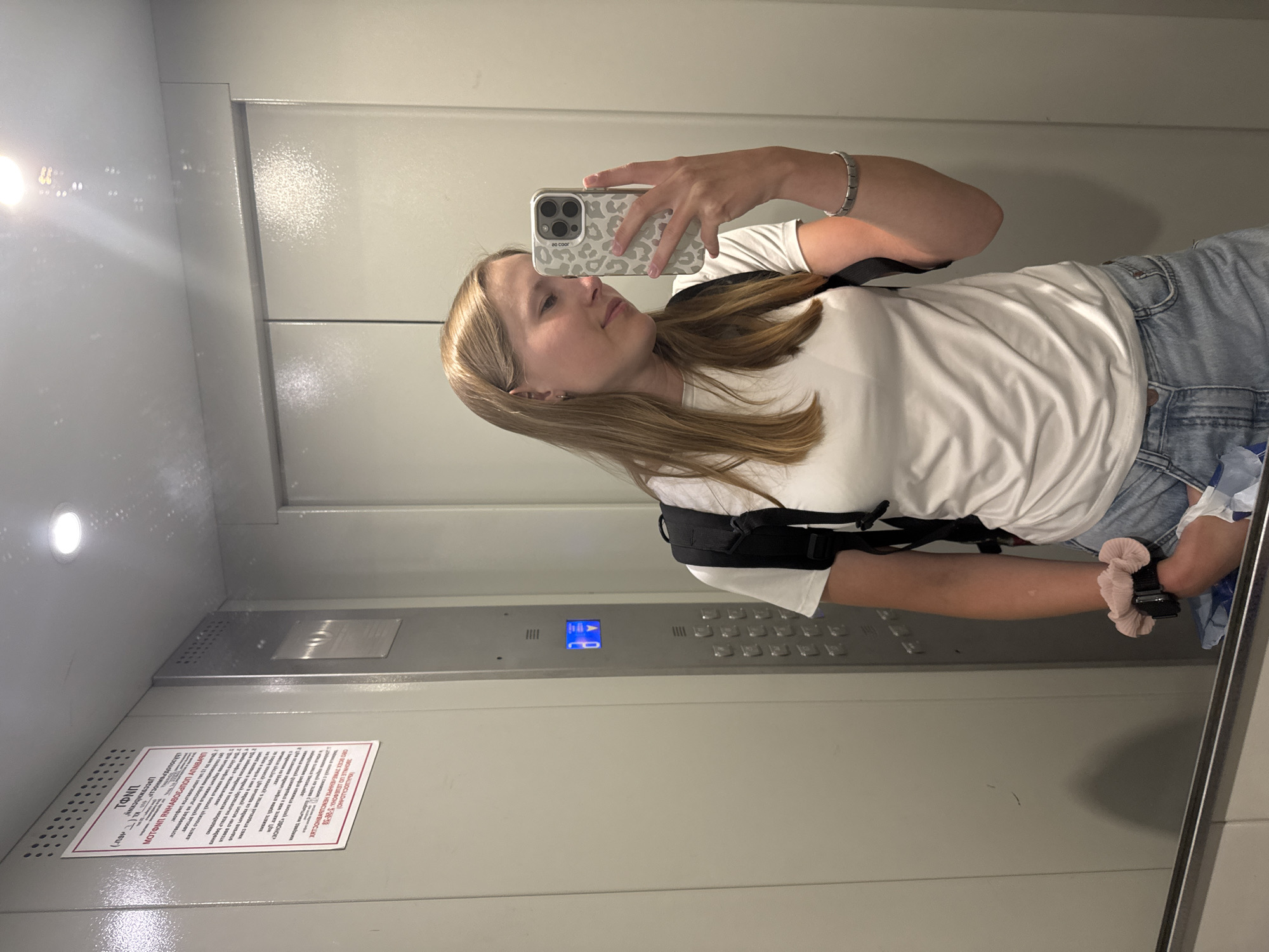 The image is a selfie taken by a fair-skinned young woman with blonde hair, wearing a white t-shirt, blue jeans, and a black backpack, in what appears to be an elevator. She is holding a smartphone with a patterned case in her right hand, using it to capture the reflection in the mirror-like elevator walls. The elevator's interior is gray, with visible buttons, a display panel showing "16", and a warning sign in Cyrillic script. A watch is visible on her left wrist, and a pink hair tie is also present. The location is indicated by the Cyrillic writing on the sign in the elevator, which suggests the image was taken in Chaikovsky, Russia.