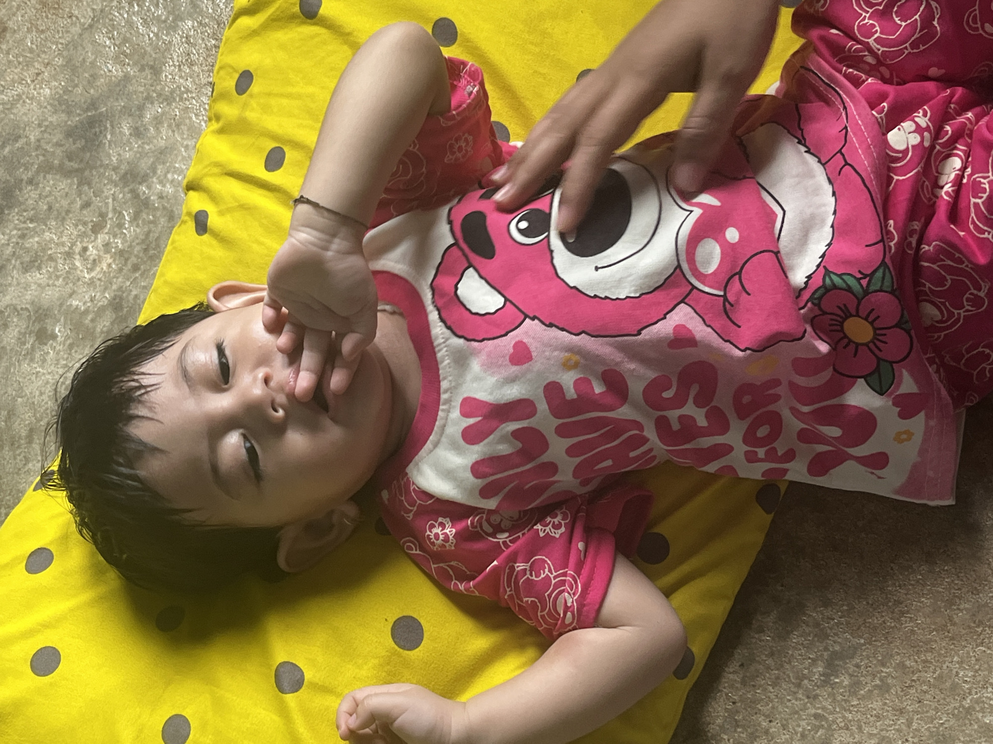 A young child, likely a toddler, is lying on their back on a yellow mat with dark grey polka dots. The child has dark hair and is wearing a pink and white t-shirt featuring a cartoon bear and text. The child's right hand is near their mouth, and their left arm is bent with the hand resting near their head. Another person's hand is visible in the foreground, touching the child's face. The background appears to be a textured, light-colored surface, possibly a floor. The scene is likely indoors, given the artificial lighting and the nature of the mat and floor. The location in Meliau Hulu, Indonesia is a residential area, as suggested by the presence of a child on a mat in a home environment.