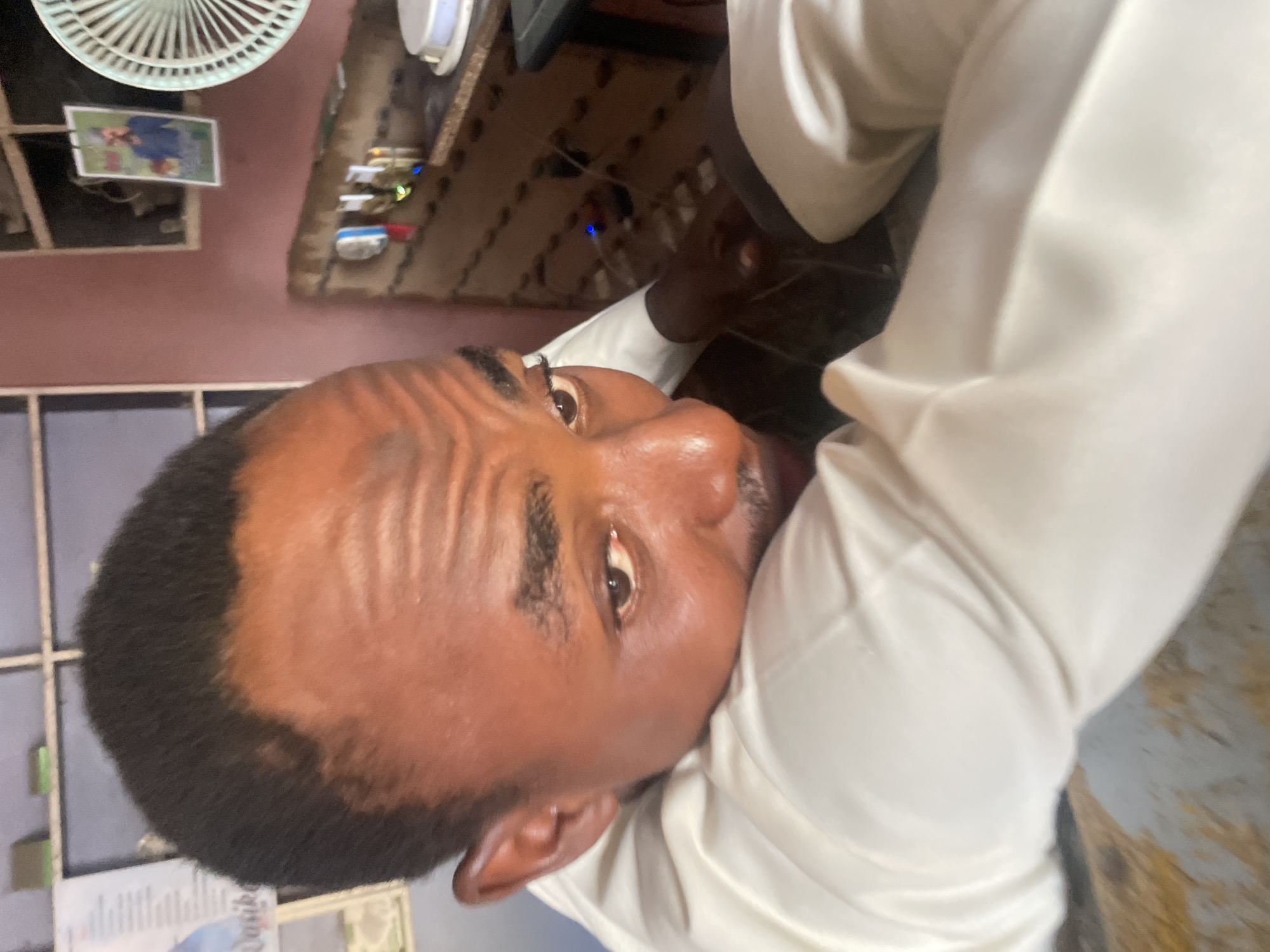A portrait-oriented image captures a Black man from the chest up, looking upwards and slightly to his right (from his perspective). He has short, dark hair, a neatly trimmed beard and mustache, and prominent wrinkles on his forehead. He is wearing a light-colored, possibly white or off-white, collared shirt. His expression appears neutral or slightly contemplative.
The setting is indoors, with a reddish-brown wall visible behind him. In the upper left background, a white ceiling fan is partially visible. Below the fan, a small, framed picture hangs on the wall, appearing to depict two blurry figures, possibly people. To the right, what seems to be a desk or shelf holds various small objects, including electronic devices indicated by tiny blue and green lights. The background elements suggest a somewhat cluttered personal space, such as an office or living area. No specific activity beyond looking upwards is clearly identifiable. The time of day and external weather are not discernible. No text is clearly readable in the image or its provided crops.
