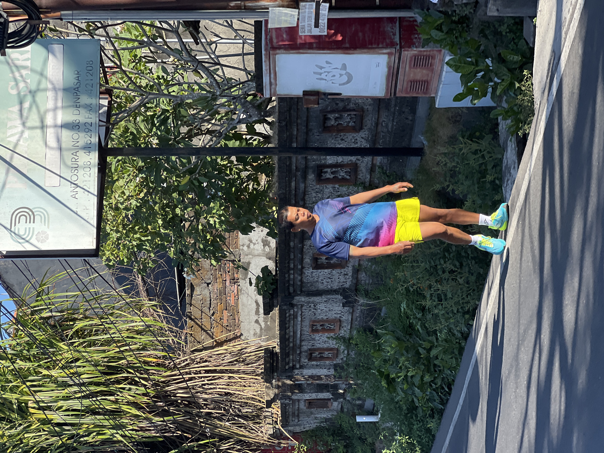 A man with dark hair walks along a paved road in Denpasar, Indonesia, casting a shadow on the asphalt. He is wearing a colorful t-shirt with diagonal blue, pink, and yellow stripes, and bright yellow shorts. His feet are clad in blue and neon green athletic shoes with white socks. The man is walking away from the viewer towards the right of the frame, with his head turned downwards.
To his left, a low stone wall, decorated with alternating square openings, runs along the edge of the property. Dense green foliage grows behind the wall. Tall, thin stalks of dry bamboo or reeds are bundled together on the left side of the image, leaning against a weathered brick and concrete wall.
To the man's right, a large, red and white utility box stands beside the road. The white panel on the front of the box features a large stenciled image of a handprint. A sign is attached to the right side of the utility box, with some text visible. Above the wall and behind the man, a portion of a teal-colored sign with white text and graphics is visible, mounted on a black pole. Text on the sign reads "ANTOSURA NO. 33 DENPASAR" and phone numbers. The environment appears sunny, with distinct shadows indicating a bright day. The overall setting suggests a street or roadside in a tropical urban area.