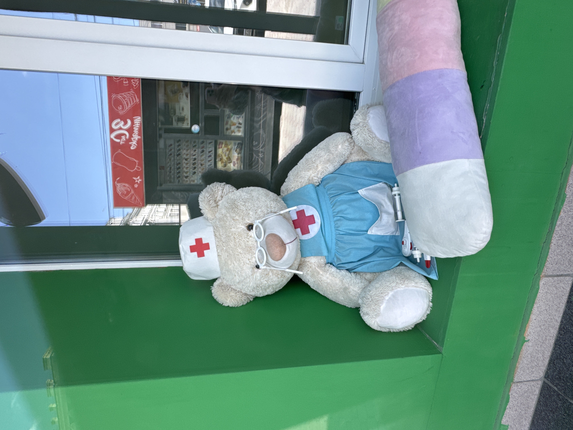An outdoor shot shows a teddy bear dressed as a medical professional, sitting on a green ledge next to a window. The teddy bear wears a white nurse's cap with a red cross, a blue uniform with a red cross emblem, and glasses. A large, cylindrical pillow with pastel stripes of white, lavender, and pink rests in front of the teddy bear. Through the window, a bright blue sky is visible, along with a red sign that appears to advertise ice cream with Cyrillic text. The reflection in the window shows buildings and possibly a street. The scene suggests a display or decoration, possibly outside a pharmacy or medical-related establishment. The context of Novosibirsk, Russia, points to the location being a city in Siberia, known for its industrial and scientific presence.