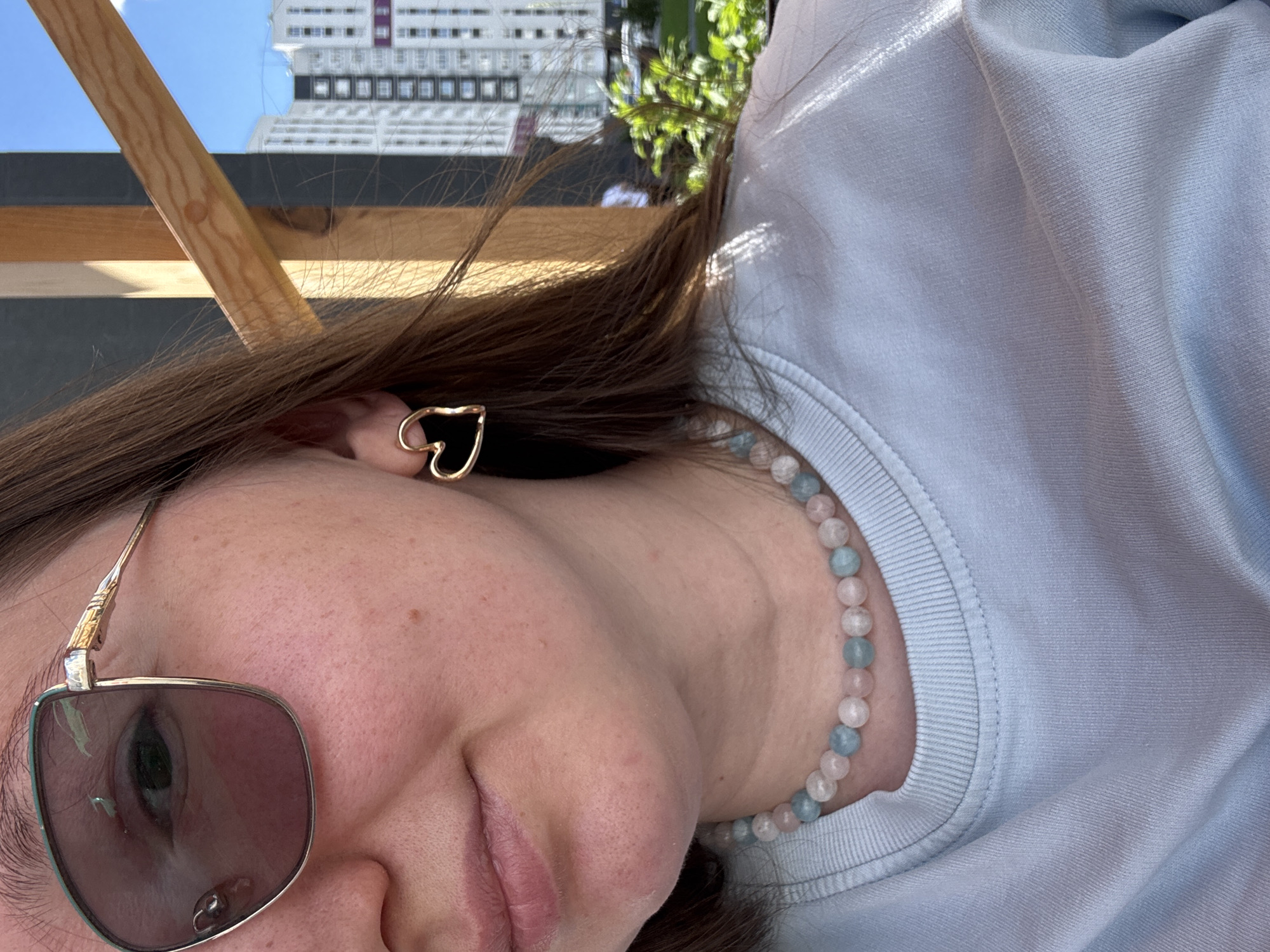 The media file shows a close-up, horizontally oriented selfie of a young woman outdoors. She is wearing sunglasses with pinkish-purple lenses and a gold heart-shaped earring. A necklace made of pale pink and blue beads is visible around her neck, and she is wearing a light blue sweatshirt. Her hair is brown and falls around her face. The background is blurred but appears to show a modern building with many windows against a clear blue sky, suggesting it is daytime with good weather. Parts of a wooden structure are also visible in the foreground, and some greenery can be seen. The overall impression is casual and relaxed. The selfie is taken from a low angle, emphasizing the woman's face and upper body. There is no visible text in the image.