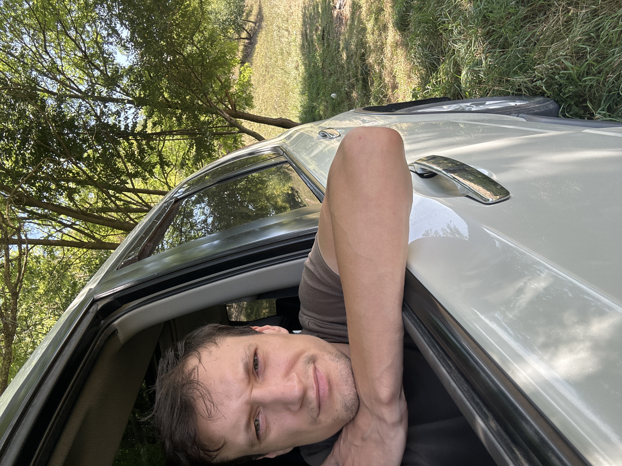 In Makiv, Ukraine, a man with short brown hair and stubble leans his forearms on the open window frame of a white vehicle. His head is slightly tilted, and he looks directly at the camera with a neutral expression. He is wearing a brown t-shirt. The vehicle's interior, including dark upholstery and a window seal, is visible. Outside the car, lush green trees and a sunlit grassy area suggest a rural or park-like setting. The car appears to be parked on a slight incline, with its right side tires visible in the tall grass. Sunlight filters through the leaves of the trees, casting dappled shadows on the ground.