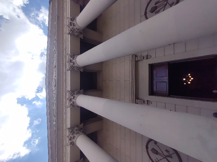 The image depicts the neoclassical facade of the Metropolitan Cathedral of Buenos Aires, located in the San Nicolás neighborhood, facing Plaza de Mayo, Buenos Aires, Argentina. The perspective is a low-angle shot, looking upwards towards the building's portico and the sky.
The central feature is a series of six large, smooth, light-colored columns with ornate Corinthian capitals, supporting an entablature. Above the capitals, a detailed frieze featuring sculpted figures is visible, topped by a prominent cornice. The wall behind the columns is constructed of light-toned masonry, exhibiting horizontal coursing.
At the base of the central columns, a large, dark wooden double-leaf door serves as the main entrance, appearing partially ajar. Through the opening, a dimly lit interior reveals a chandelier with multiple glowing bulbs. To the left and right of the entrance, on the lower wall section, circular relief sculptures are present; the one on the left appears to depict crossed implements or symbols.
The upper portion of the image shows a blue sky with scattered white cumulus clouds. A dark, indistinct form, consistent with the top of a head or shoulder, is partially visible at the very bottom center edge of the frame, indicating the vantage point is close to the building's entrance. The scene is illuminated by natural daylight. No distinct individuals are clearly visible or engaged in specific actions.