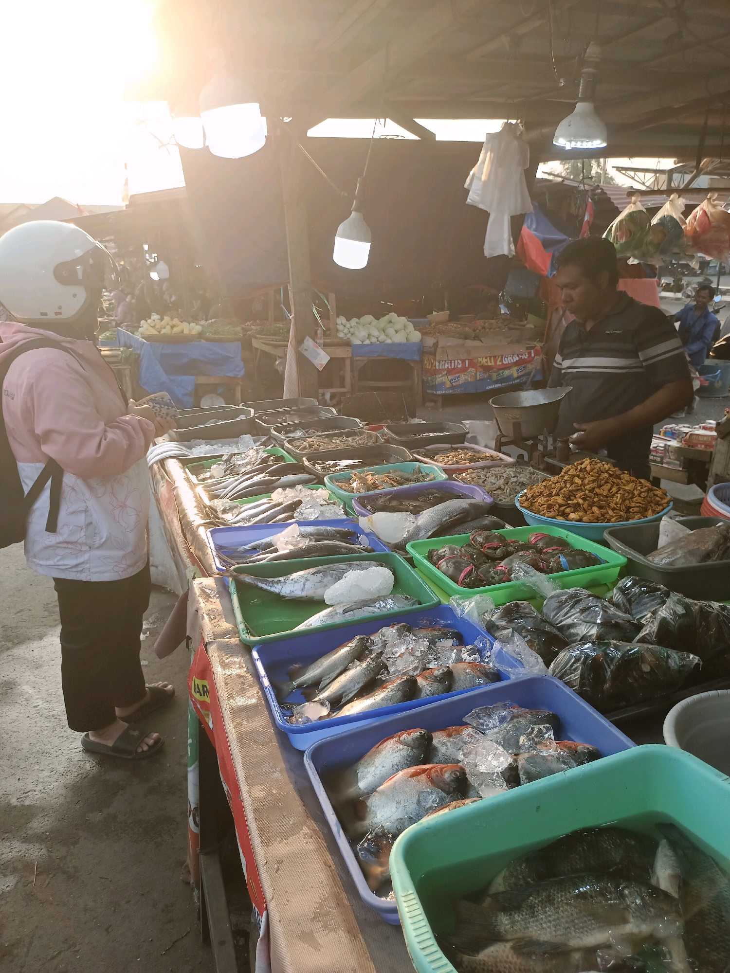 This image captures a bustling scene at a fish market, likely in Karawang, Indonesia. The sun is shining brightly, casting a warm glow over the market stalls. In the foreground, a person wearing a pink jacket and a white helmet stands looking at the fresh produce displayed on a table. The table is laden with various types of fish, crabs, and other seafood, arranged in plastic containers filled with ice. A vendor, a man wearing a striped shirt, stands behind the table, attending to his customers. The market appears to be open-air, with a temporary roof structure overhead providing shade. Bags of merchandise hang from the ceiling, adding to the lively atmosphere of the market. The ground is a dirt surface, typical of many outdoor markets in the region. The overall impression is one of a vibrant and active marketplace, full of fresh goods and local commerce.
