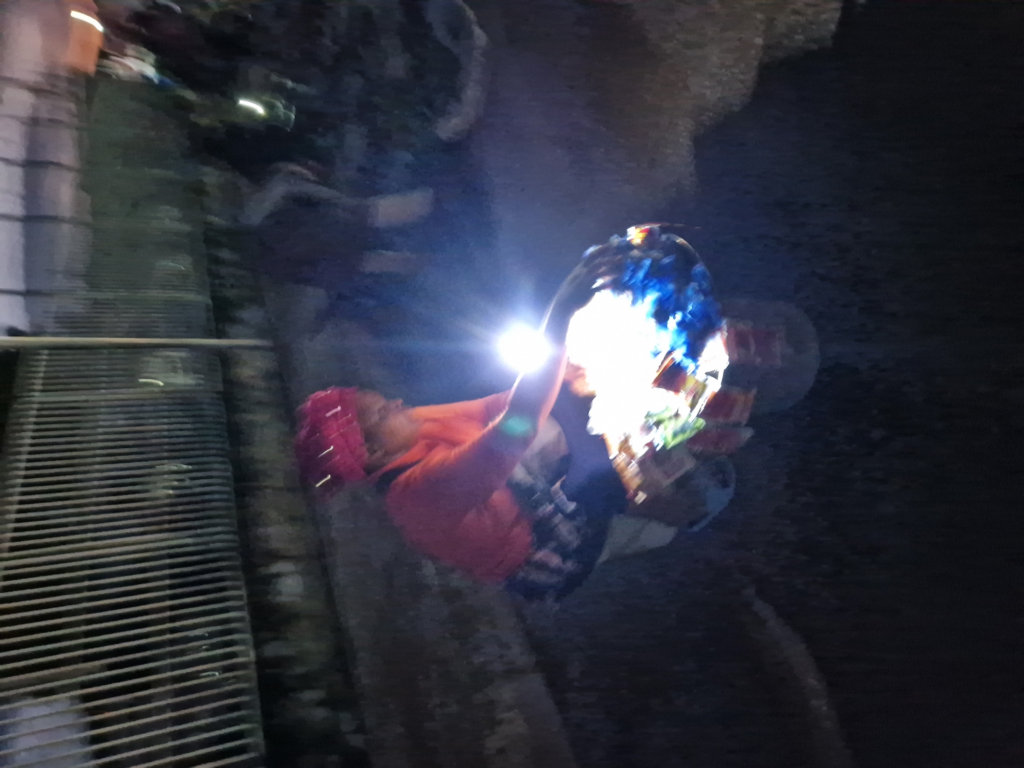 A person wearing a red headscarf and an orange shirt is seated at a low table displaying various items. The person is using a bright light source, possibly a flashlight, to illuminate the items on the table, which appear to be packaged goods. The scene is set outdoors at night, with a metal fence visible in the background. The ground is dark asphalt. The image is blurry due to low light conditions and motion. The location is stated to be Kabong, Nigeria.