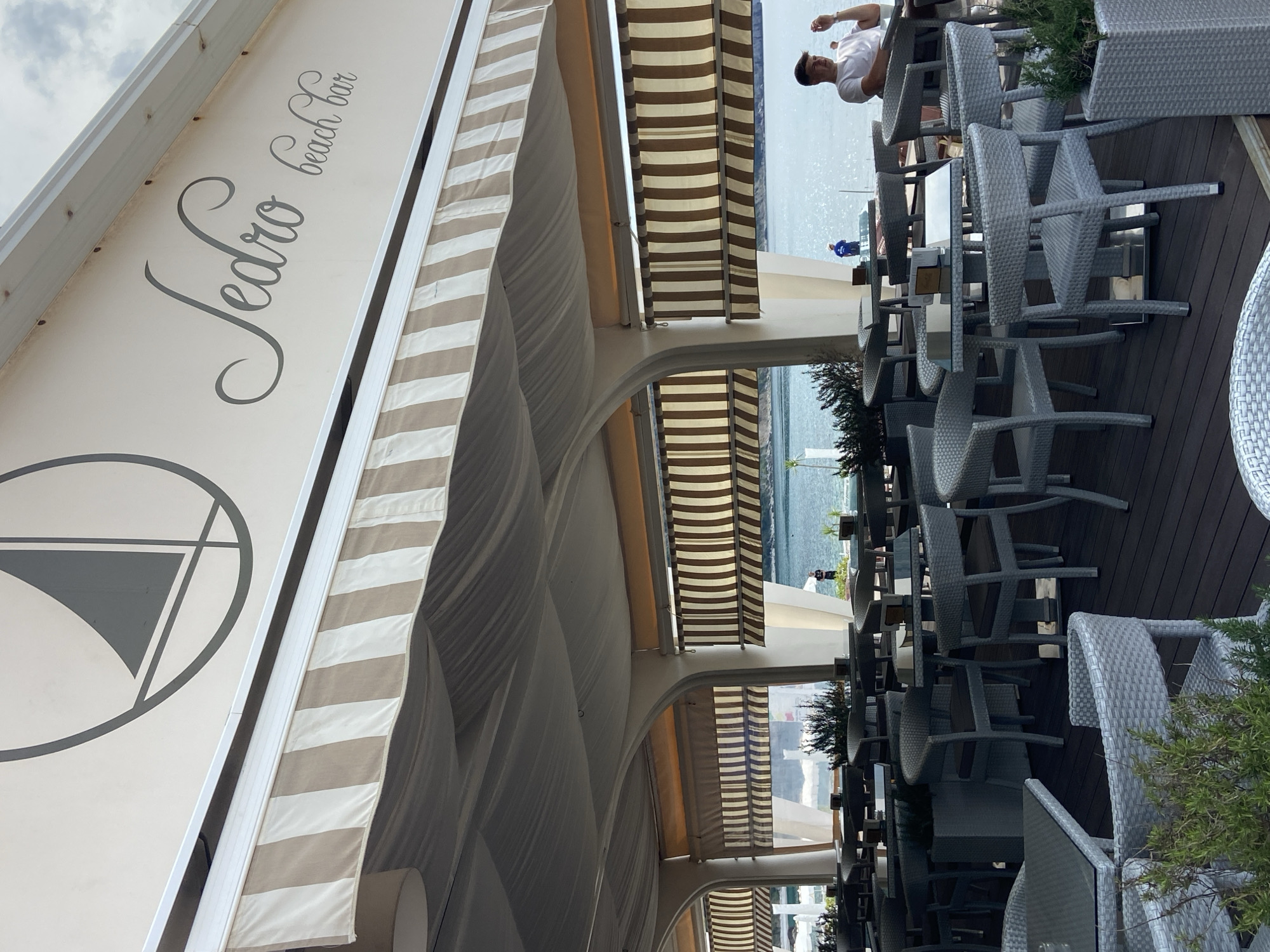 The image depicts the outdoor seating area of "Jedro beach bar," featuring rows of tables and chairs under a striped awning. A man wearing a white shirt and dark hair is seated at a table towards the back. The background shows a body of water, likely the sea, with people visible in the distance. The location is Prijevor, Montenegro, as indicated by the name of the bar. The chairs are made of woven material in a gray hue, and some tables have plants as centerpieces. The decking is made of dark wood. The weather appears overcast based on the sky visible in the upper left corner.