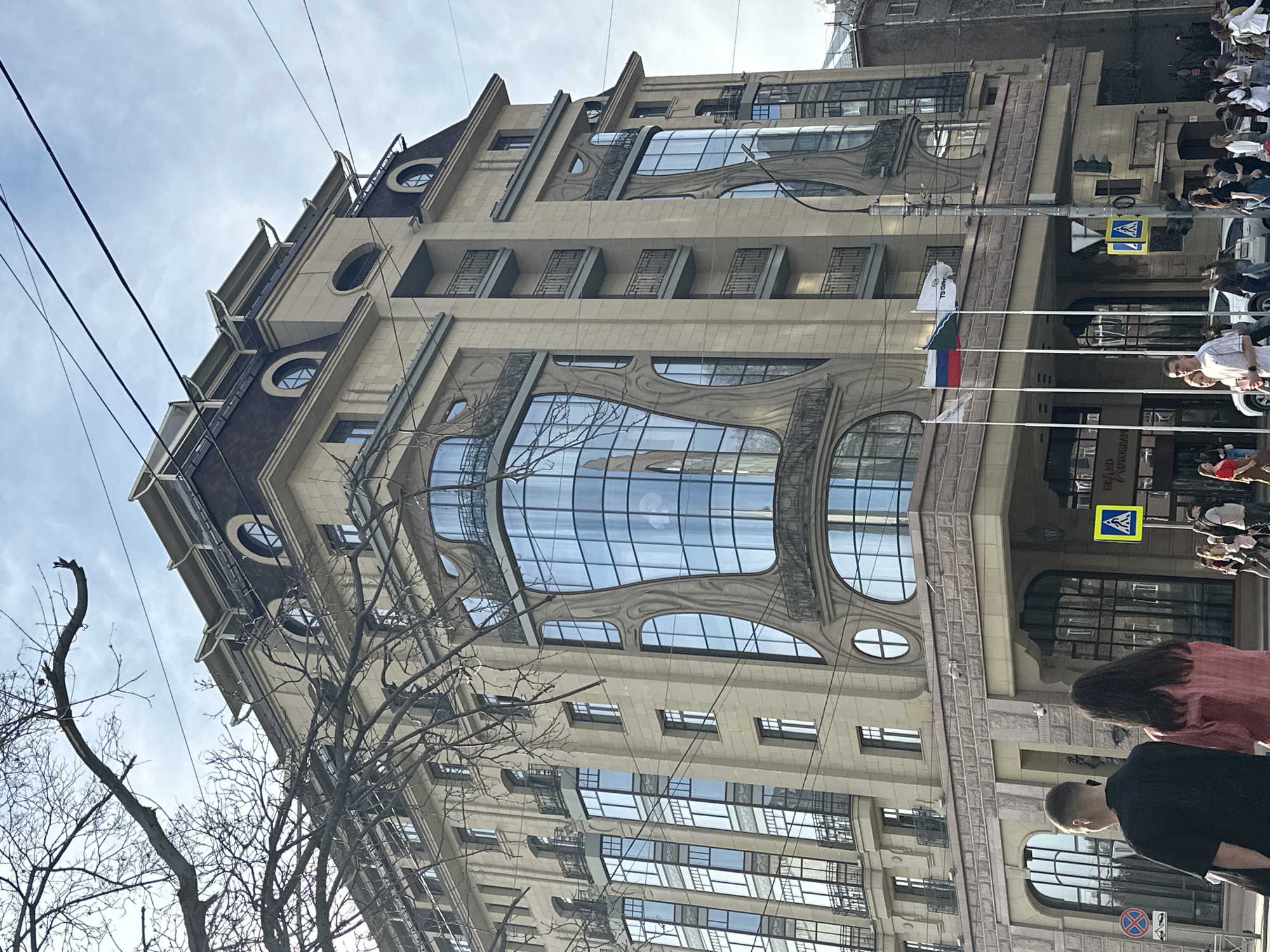 The image captures the façade of the Novosibirsk State Art Museum in Novosibirsk, Russia. The building exhibits an architectural style with large, arched windows and decorative elements. The Russian flag and other flags are prominently displayed near the entrance. Pedestrians are walking along the sidewalk in front of the museum. Bare tree branches are visible in the foreground, suggesting it could be late autumn or early spring. Overhead power lines are also visible against a partly cloudy sky.
