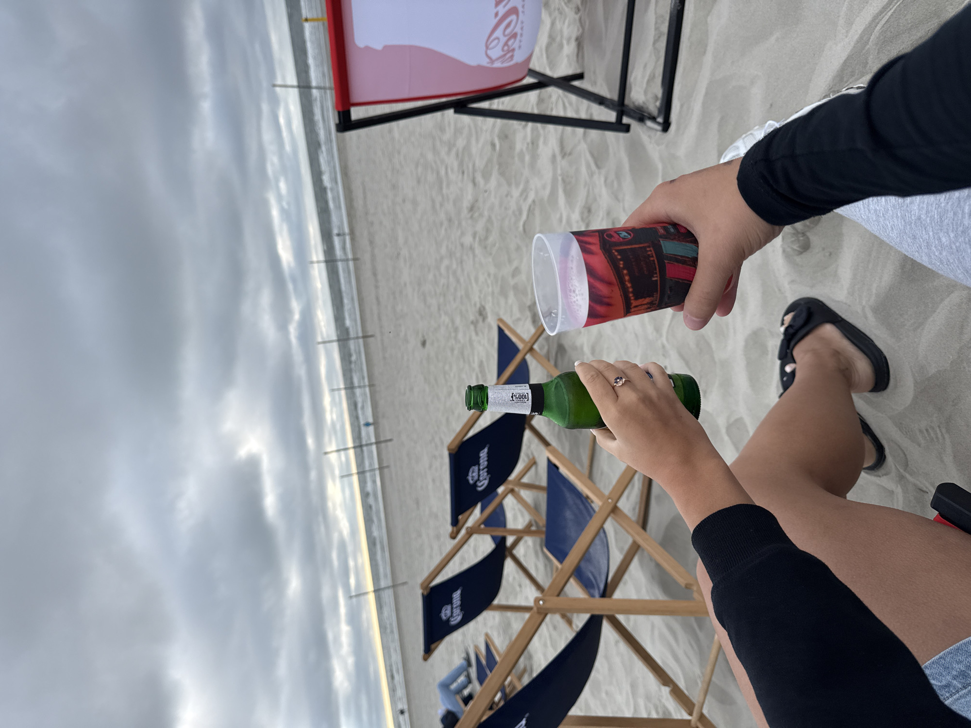 In Langeoog, Germany, two individuals are enjoying beverages on a sandy beach under a cloudy sky. The foreground shows their hands raised, clinking a green glass bottle of Corona beer and a plastic cup filled with a reddish beverage. The individuals are seated, with one person wearing denim shorts and a black long-sleeved top, and the other person wearing grey leggings and a black long-sleeved top. In the background, several navy blue beach chairs with the Corona logo are arranged on the sand. Further in the distance, the ocean waves are visible, and a line of tall, thin posts stretches across the beach. To the right, a red and white beach chair with a Coca-Cola logo is partially visible. The overall scene depicts a relaxed moment of enjoyment on a German beach.