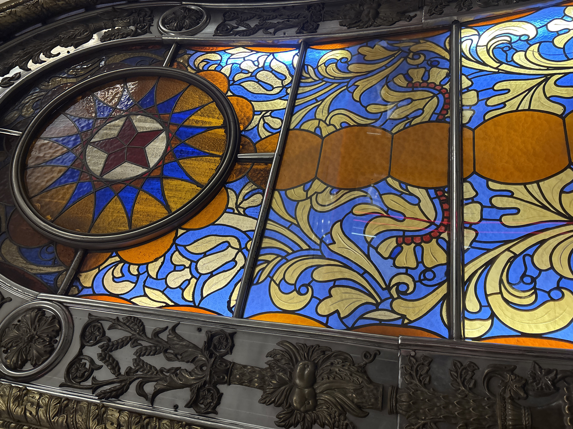 This is a close-up photograph of a stained-glass window with elaborate metal detailing. The window contains a circular design on the left featuring a red star surrounded by geometric patterns in blue, gold, and brown. To the right of the circle, the window displays ornate floral and flowing designs in blue, gold, and brown. Vertical metal bars divide sections of the stained glass. The metal frame and adjacent architectural details below the window are decorated with floral and stylized motifs. Based on the star emblem and the architectural style, the location is likely within the Moscow Metro, Russia, specifically a station dating from the Soviet era.