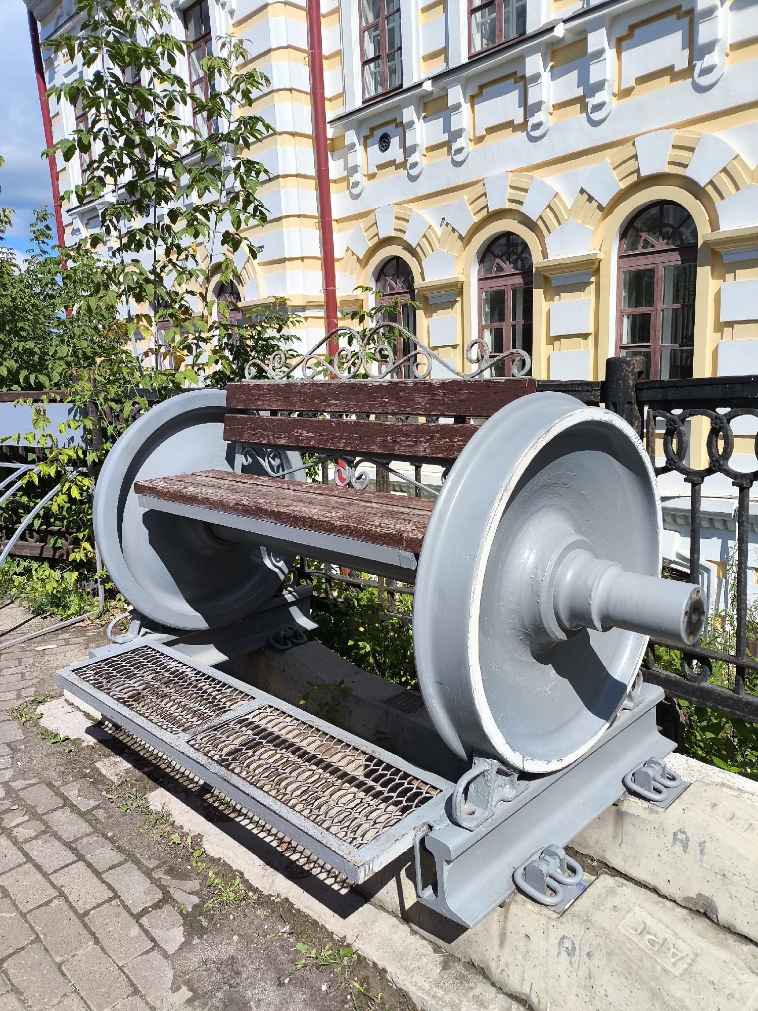 This image shows a unique park bench constructed from two large, grey train wheels. The bench has a dark brown wooden seat and backrest, with decorative metalwork supporting the back. In front of the bench is a metal grate platform. The bench is situated outdoors on what appears to be a paved walkway, with greenery and a building visible in the background. The building is light-colored with yellow decorative accents and arched windows. This is the train wheel bench located in the Square of the Soviet Air Fleet in Perm, Russia.