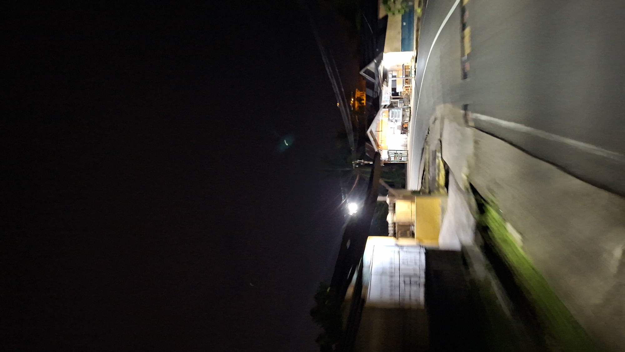 The image depicts a street scene at night in Kutaglumpang, Indonesia. The perspective is low and angled, suggesting the photo was taken from a moving vehicle or at ground level. On the left side of the frame, a blurred, light-colored rectangular structure is visible, possibly a building or a wall, with some green foliage and grass to its left. To the right of this structure, a yellow concrete barrier or wall is present.
Further down the street, several illuminated buildings are visible. These appear to be small businesses or shops, with bright interior lighting spilling out. One building is particularly prominent, with a white facade and visible tables and chairs inside. There are also power lines strung across the scene.
The road surface is dark asphalt, with white lane markings visible. There are speed bumps present on the road. The sky above is dark and appears to be night, with no distinct stars visible. There is a faint, curved light streak in the upper right portion of the sky, and another faint greenish arc in the upper center. The overall impression is that of a quiet, possibly rural or semi-urban street at night, with commercial activity present in the illuminated buildings.