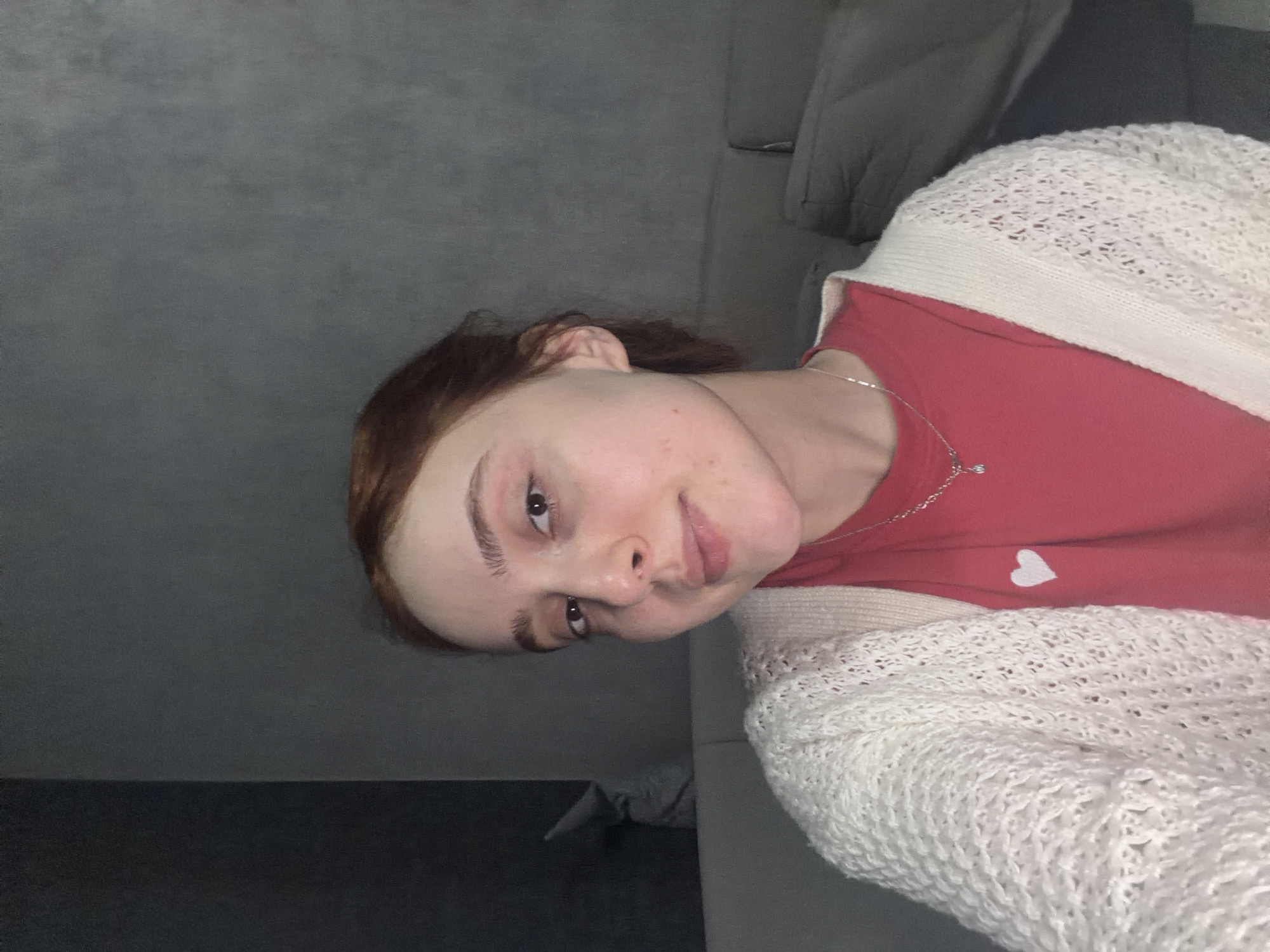 A young woman is depicted in a close-up, casual portrait, rotated 90 degrees clockwise. She has reddish-brown hair pulled back from her face and dark eyebrows and eyes. Her skin appears fair with a few minor blemishes or freckles visible. She is looking directly towards the camera with a slight smile or smirk.
She is wearing a bright red top, on the right side of which a small white heart emblem is visible. Over the red top, she has a light-colored, possibly cream or off-white, open-knit cardigan with a distinct textured pattern. A delicate silver chain necklace with a small charm hangs around her neck.
The background suggests an indoor setting, likely a living space. A plain gray wall takes up the upper left portion of the image (when oriented correctly), and a darker gray sofa or upholstered furniture is visible behind her, particularly on the right side. The overall lighting is soft, indicative of an indoor environment without strong direct light. There is no visible text in the image. The mood is casual and relaxed.