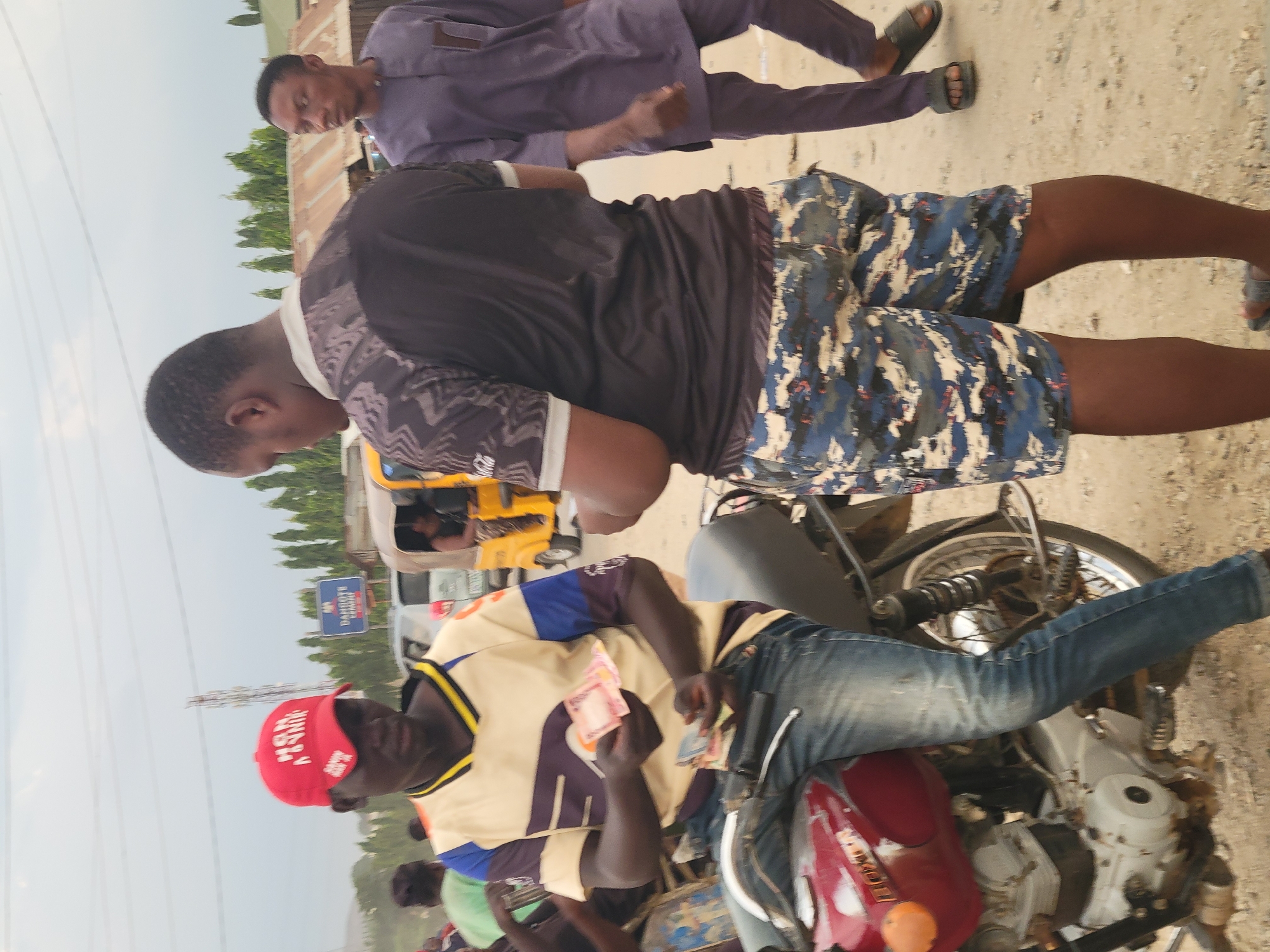 In Madalla, Nigeria, a man in a red cap, cream and brown jersey, and blue jeans sits on a parked red motorcycle, counting currency. Two other men stand nearby, one in a purple outfit and the other in a black shirt and blue camouflage shorts. In the background is a yellow tricycle and a sign that reads "DANGIWA SCHOOL." The environment appears to be a roadside or marketplace.