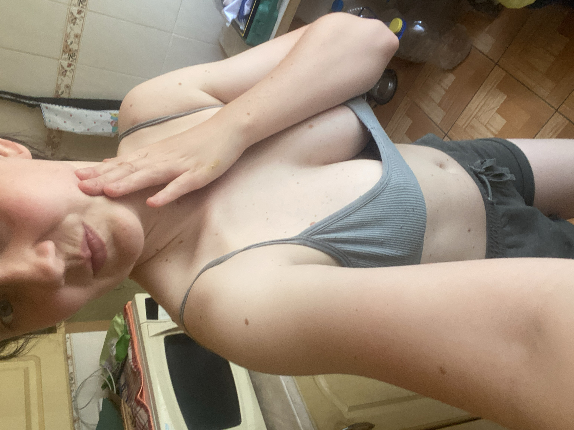 A woman is shown in a selfie-style image, likely taken indoors given the tiled wall and cabinetry visible in the background. She is wearing a grey ribbed tank top and dark shorts. Her right arm is raised, with her hand resting on her neck, and her left arm is crossed over her chest. Her head is tilted downwards, and her gaze is directed towards the camera. The lighting appears to be artificial, suggesting it could be any time of day. There is no visible text in the image. The overall mood is casual and intimate, with the woman appearing relaxed and confident. The background shows elements of a kitchen or bathroom, with tiles on the wall and wooden flooring. There are also some items on shelves, including what appears to be a bottle of water. The image is an artistic representation of the subject, focusing on her pose and attire rather than a specific event or detailed setting.