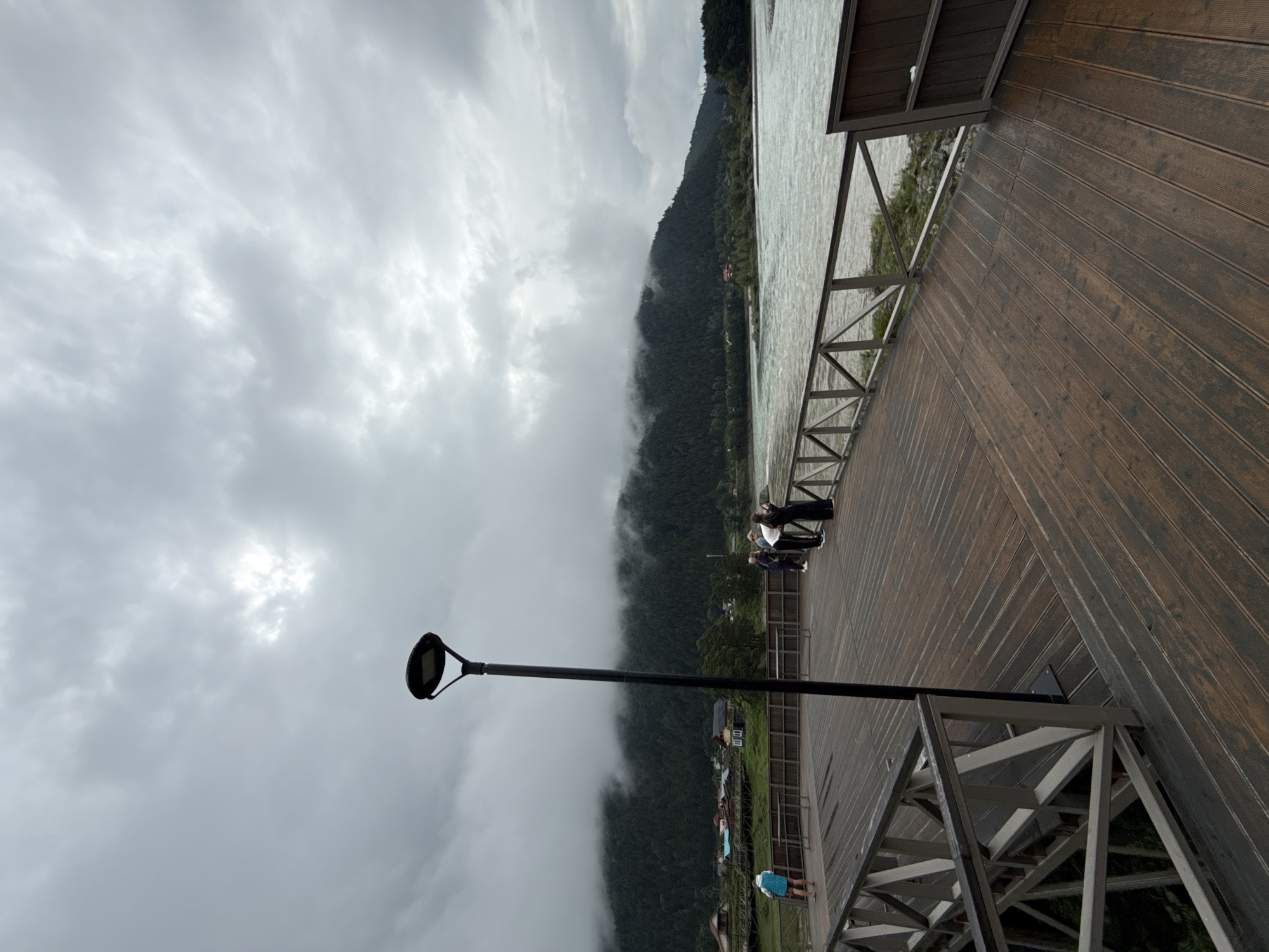 This is a vertical, eye-level, full shot of a wooden pier overlooking a wide, choppy river under a cloudy sky. The pier is made of dark, weathered wood planks. A black lamppost stands tall in the center-left of the frame. Several people are scattered across the pier: on the far left, a person in a blue jacket stands with their back to the camera, facing the river. Further to the right, a group of three people are gathered, two appearing to be looking at a phone. In the background, a dense, dark green forest covers a mountain range, with low-hanging clouds partially obscuring the peaks. Several small buildings and wooden structures are visible nestled at the base of the mountains. The location is Elekmonar, Russia.