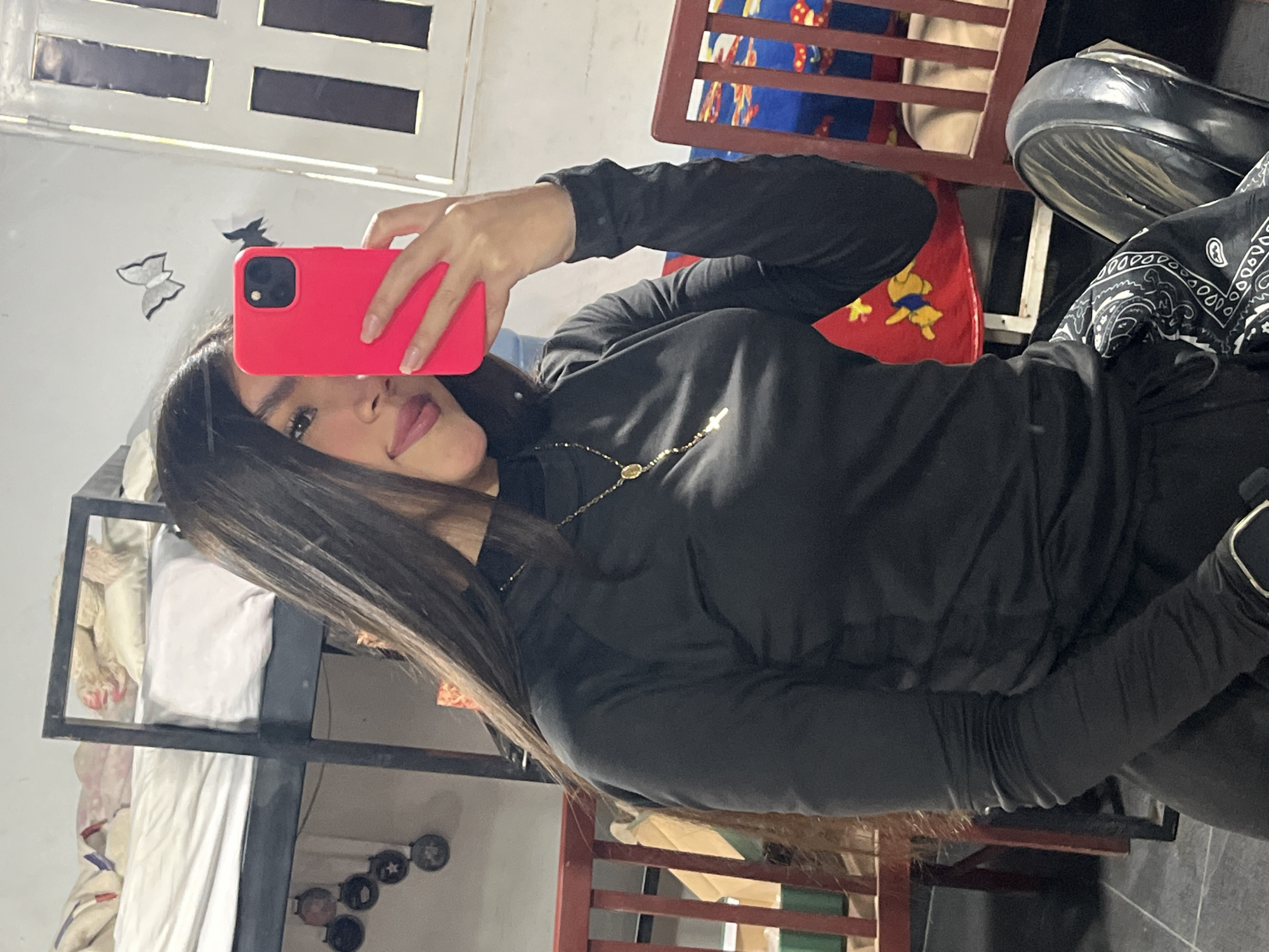 A young woman, partially visible in a mirror selfie, is seated indoors in what appears to be a residential setting in Juan Griego, Venezuela. She has long, dark hair, light brown skin, and is wearing a black long-sleeved turtleneck top with a gold-colored rosary-style necklace featuring a cross pendant. Her lips are adorned with dark pink lipstick, and her fingernails have a light pink polish. She holds a bright pink smartphone with a visible black camera module in her right hand to capture the reflection. A black smartwatch with a silver bezel is present on her left wrist. She is wearing dark pants, and a black and white paisley-patterned fabric is visible on her lap. She is seated on a black chair with a polished backrest.
The background reveals a sparsely furnished room. To the left, a dark metal bunk bed is present; the top bunk contains a patterned blanket and a white pillow, while the bottom bunk has white bedding. Below the top bunk, five circular decorative objects or medallions are affixed diagonally to the wall. To the right, a white window frame with multiple vertical panes is visible. Two butterfly-shaped wall decorations, one white and one black, are mounted on the wall near the window. A brown wooden chair with vertical slats and a blue cushion adorned with colorful cartoon characters (resembling Winnie the Pooh designs) is partially visible to the right. The floor consists of dark gray square tiles.
