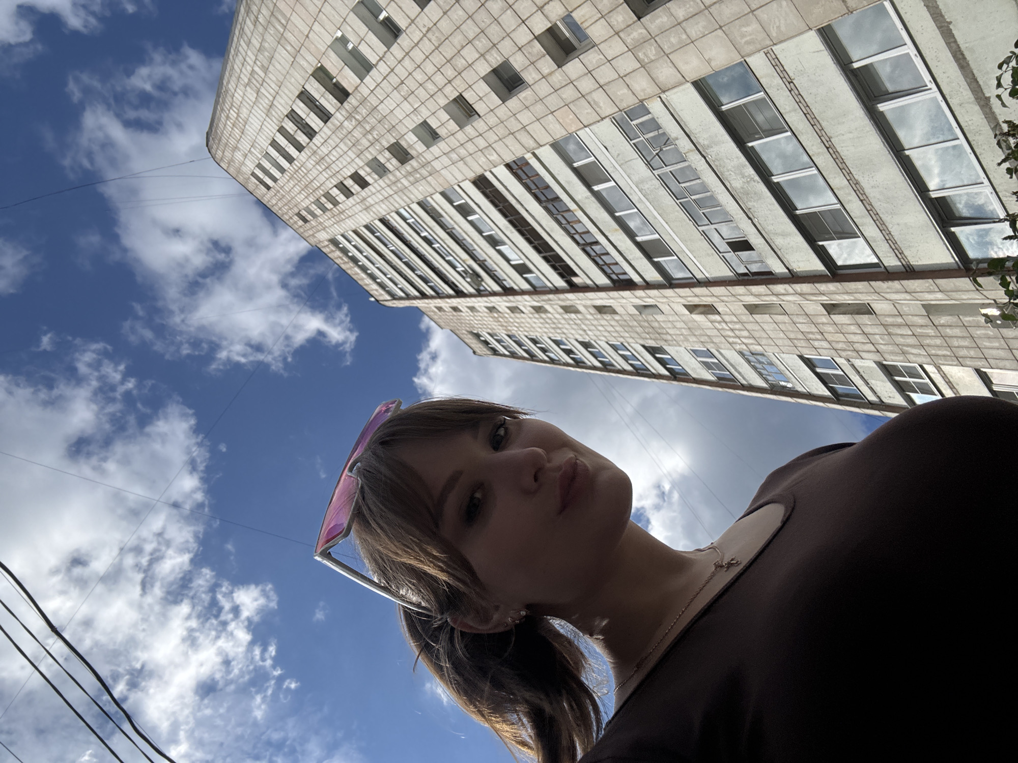 This media file is a low-angle, Dutch tilt photograph taken outdoors. The main subject is a young woman with brown hair, wearing pink-tinted sunglasses on her head and a dark brown top. She is looking up towards the camera with a slight smile.
The background is dominated by a large, modern concrete building with many windows, viewed from below. The building's architecture suggests a Soviet-era design. The sky above is blue with scattered white clouds. Several dark wires are visible crisscrossing the sky in the foreground, possibly utility lines. The lighting suggests it is daytime, likely midday given the brightness. The overall mood is casual and relaxed.
The image appears to be a selfie, captured from a low perspective looking upwards. The woman's pose and expression, combined with the upward angle of the buildings, create a sense of looking up at a grand structure. No visible text is present in the image. The provided location context is Chaikovsky, Russia.