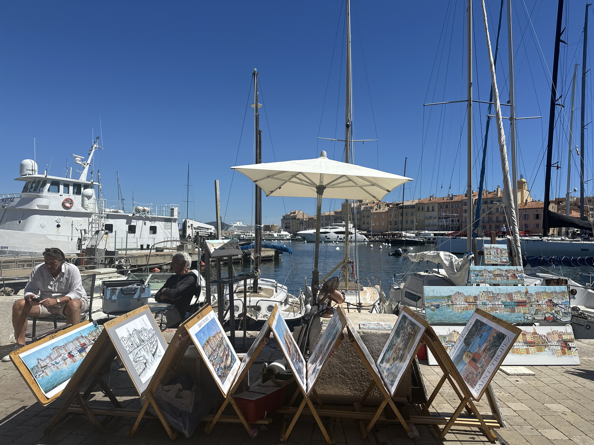 This image depicts a waterfront scene in the city of Saint-Tropez, France. The foreground features several easels displaying paintings of the harbor and its colorful buildings. Two men are seated in the middle ground, one looking at his phone and the other with his arms crossed. Behind them, a large white boat is docked, and in the background, sailboats and other vessels fill the harbor under a clear blue sky. The architecture of the buildings lining the waterfront is characteristic of the French Riviera. The overall atmosphere is one of a sunny day at a bustling Mediterranean port.