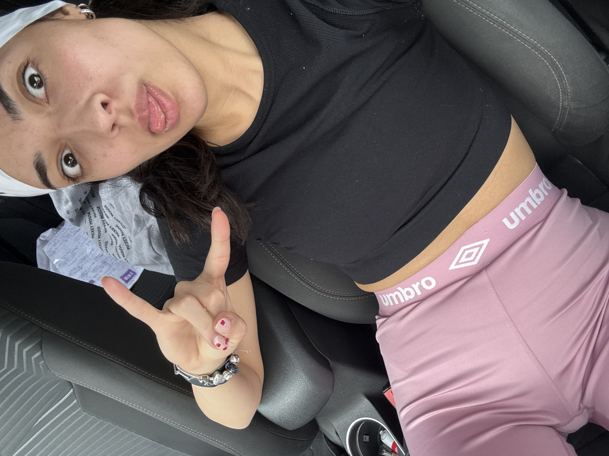 The image depicts a young woman posing for a selfie in what appears to be the interior of a car. She is wearing a black cropped t-shirt and pink leggings with the "Umbro" logo on the waistband. She has a white headband on and is making a peace sign with her hand. The seats are grey, and there are some papers in the background near her arm, as well as a bracelet on her wrist. The cup holder is visible in the frame. The image does not provide sufficient information to determine if it was taken in Sofia, Bulgaria, or to provide information about the actions, interactions, or environment.