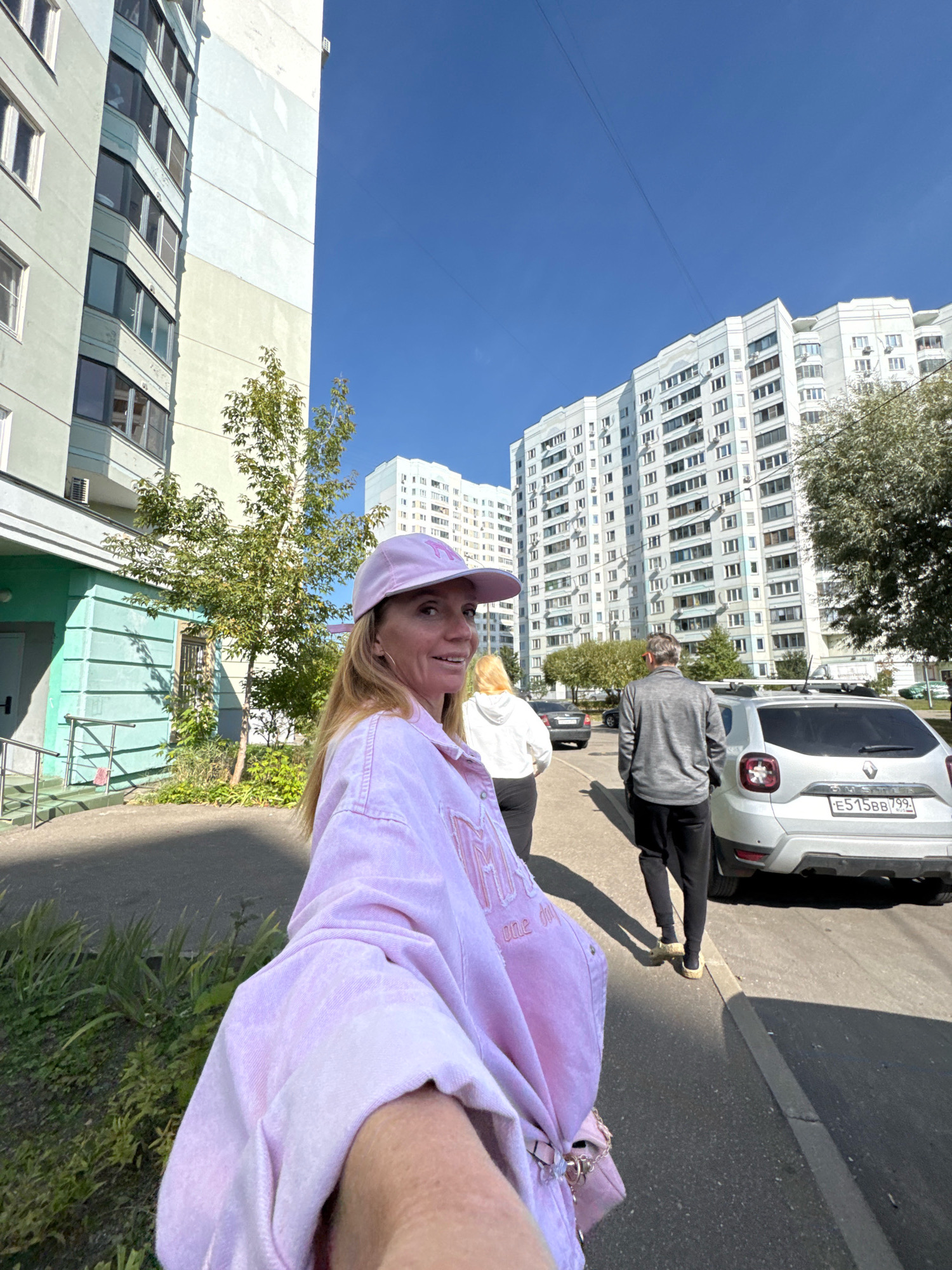 In Милицейский посёлок, Russia, on a bright, sunny day, a woman wearing a pink baseball cap and a matching pink denim jacket is captured in a selfie-style shot. She is looking over her shoulder towards the camera with a slight smile. Behind her, a woman in a white hoodie and black pants walks away, followed by a man in a grey long-sleeved shirt and black sweatpants. The group is walking on a paved sidewalk next to a street where a silver Dacia Duster is parked. The license plate of the Dacia reads "E515BB 799". The background features tall, multi-story apartment buildings, one on the left with a pale green and cream facade, and a larger white building on the right, characteristic of urban residential areas. Trees and some greenery are visible between the buildings and along the sidewalk. The scene appears to be an outdoor public space in a residential neighborhood.