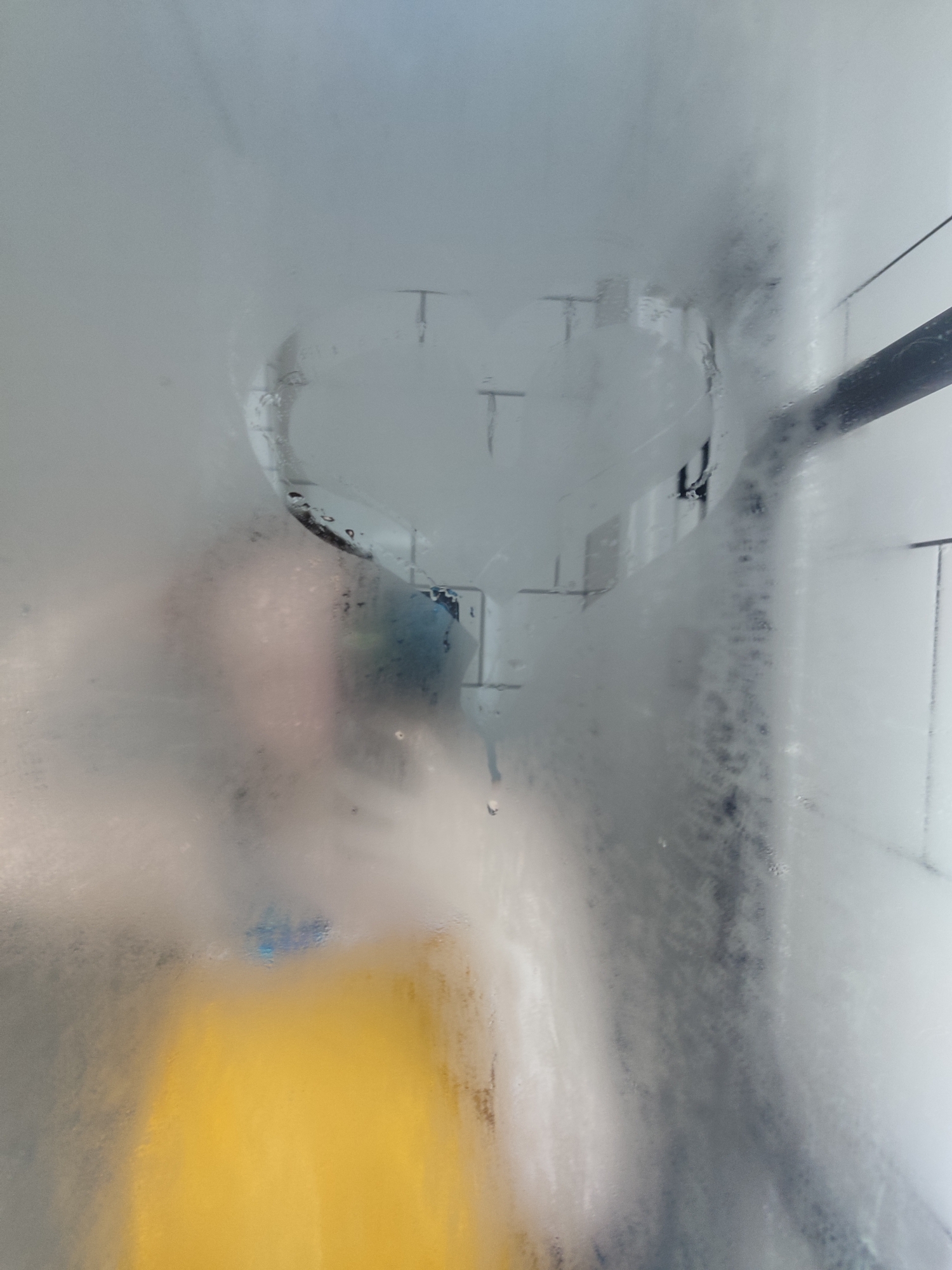 The image shows a steamy shower door with a heart drawn on it. A person, wearing a yellow garment, is visible through the steam on the left side of the frame. The background suggests a tiled shower enclosure. The location in Chaikovsky, Russia is not discernible from the visual information provided in the image.