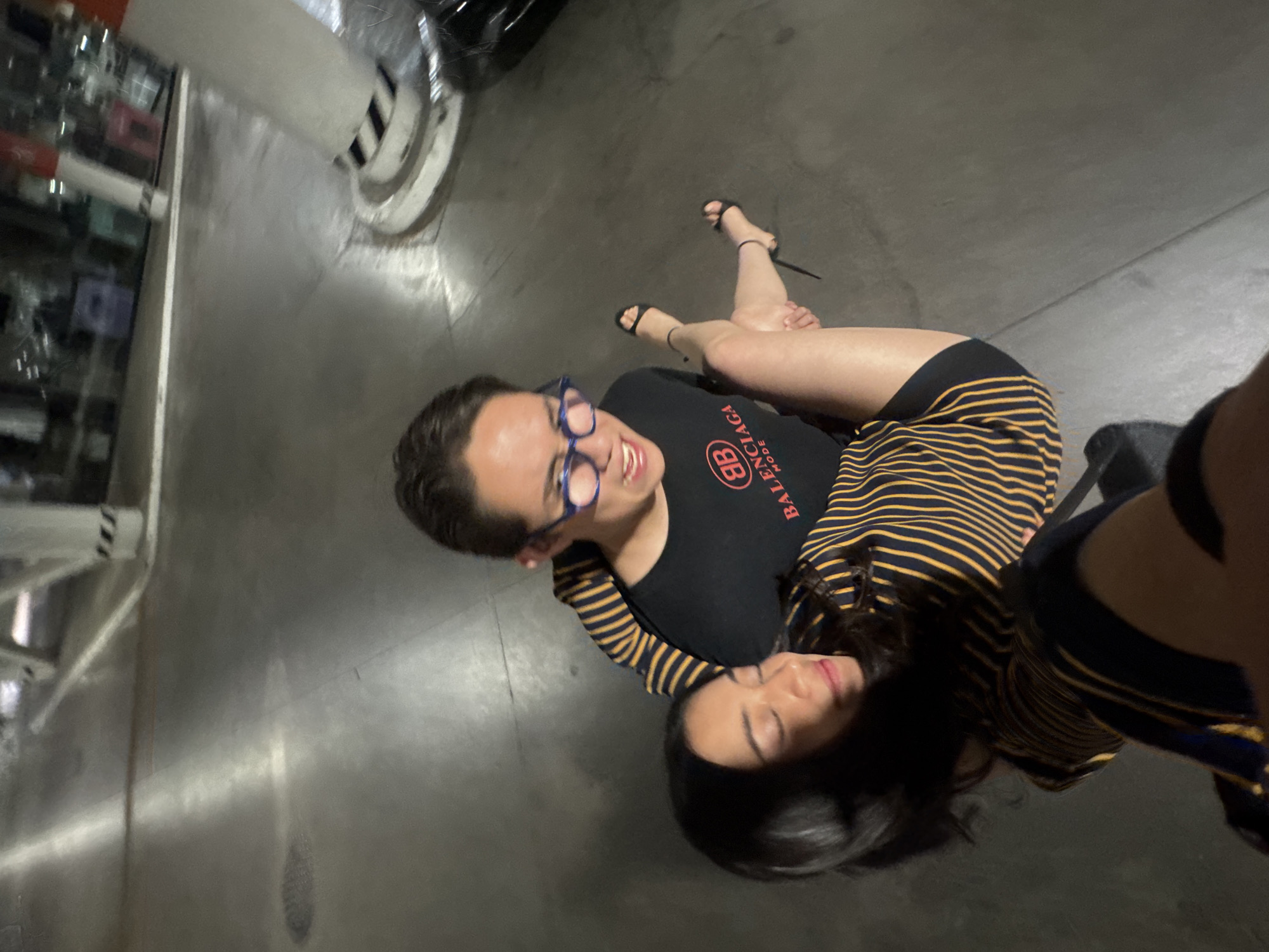 This image, captured in Puebla, Mexico, features two individuals posing in what appears to be an indoor or covered space, possibly a parking garage or industrial-style setting, characterized by concrete flooring and walls, and metallic structures visible in the background. The lighting is artificial.
One person, with short dark hair and blue-rimmed glasses, is wearing a black t-shirt that clearly displays "BALENCIAGA" and "MODE" in red text. They are smiling broadly at the camera. Their arm is around the waist of the second person, who is leaning back in their embrace. This second individual has long, dark hair, is wearing a black and yellow striped top, and black high-heeled sandals on their feet. Their eyes are mostly closed, and their expression appears relaxed and content.
The activity depicts a playful or affectionate interaction between the two, with one holding the other in a dynamic pose. The overall mood is cheerful, stemming from the visible smile and the intimate interaction.
