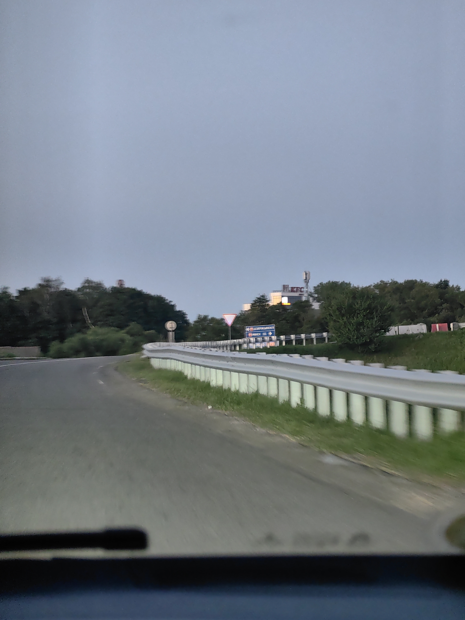 The image depicts a view from a vehicle traveling on a road, likely an exit or entrance to a highway, near the city of Fanipal, Belarus. The sky is overcast, suggesting either early morning or late evening. The road curves to the left, with a white metal guardrail running along the right side. Beyond the guardrail, a grassy embankment is visible, dotted with small white concrete barriers. In the background, a dense line of trees forms the edge of a forest. Further in the distance, a building with a prominent "KFC" sign and communication towers are visible. Road signs indicating directions to "ДЗЯРЖЫНСКІ" and "МІНСК" are also present, confirming the location within the Fanipal agricultural council region of Belarus. The foreground shows the interior of the vehicle, with the top of the dashboard and windshield wiper visible.