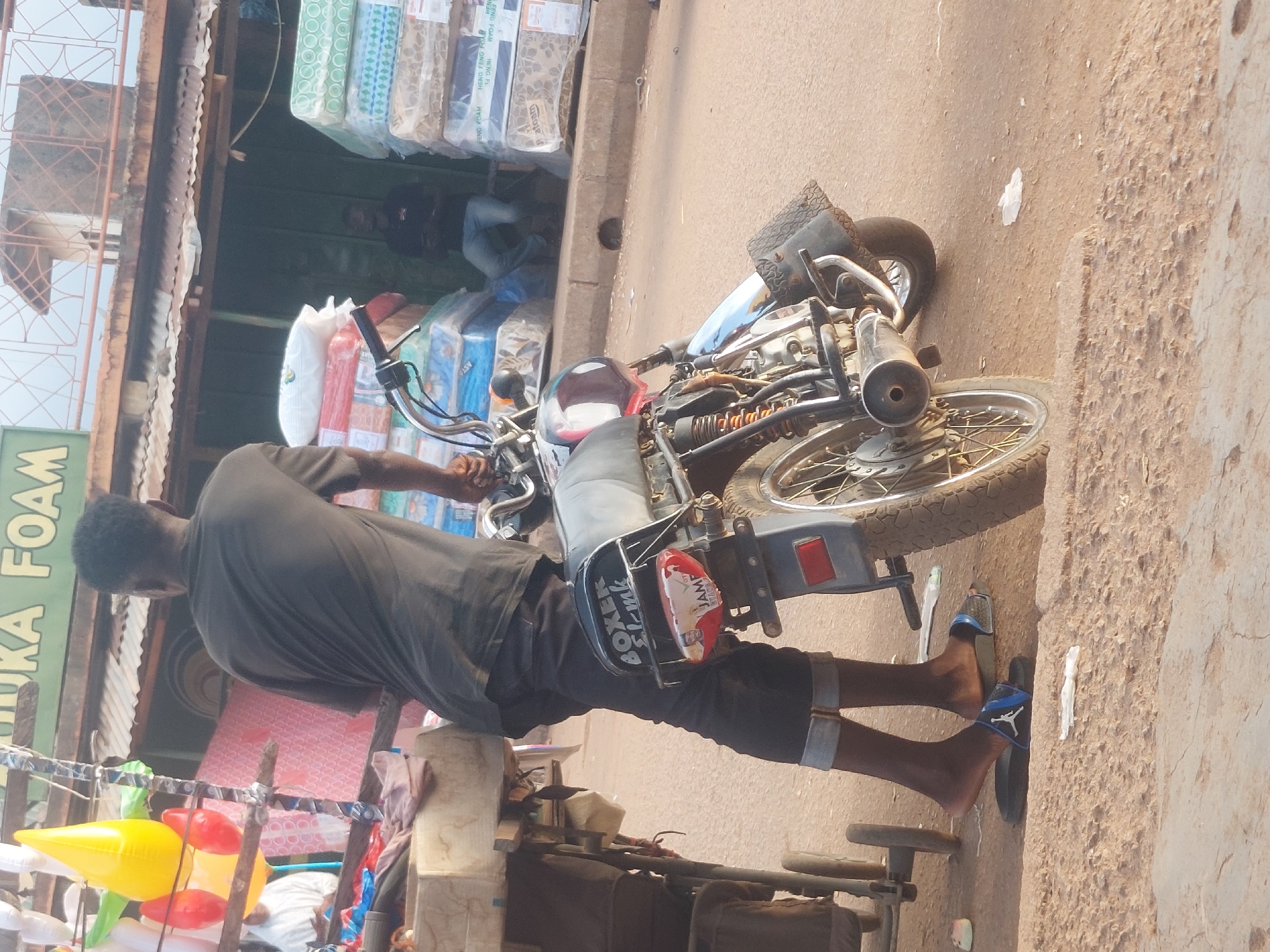This image captures an outdoor commercial scene in the city of Unknown, Unknown, during daylight hours. In the foreground, a dark-skinned male, viewed from behind, wears a dark grey short-sleeved t-shirt and dark trousers rolled at the ankles, with blue and black slide sandals on his feet. He stands beside a black "Boxer" model motorcycle, which is positioned facing right with its exhaust pipe visible. A political sticker featuring a man's face and the text "JAMES PEACE" is affixed near the motorcycle's red taillight. To the left, a rudimentary street stall displays various inflatable items, including a prominent yellow tear-drop shape and several smaller red oblong shapes. A light-colored cart or trolley is partially visible next to the stall. In the mid-ground, a store entrance is stacked high with numerous plastic-wrapped mattresses, some bearing the label "HENG FENG FOAM". A dark-skinned male is seated within this store entrance, partially obscured by the mattress stacks. Above the mattress store, a building with a corrugated metal roof and a distinctive reddish-brown metal railing or balcony is visible. The ground consists of uneven, light brown dirt and gravel, with a paved curb line further back. A small, dark object is discernible near the curb in front of the mattress store.