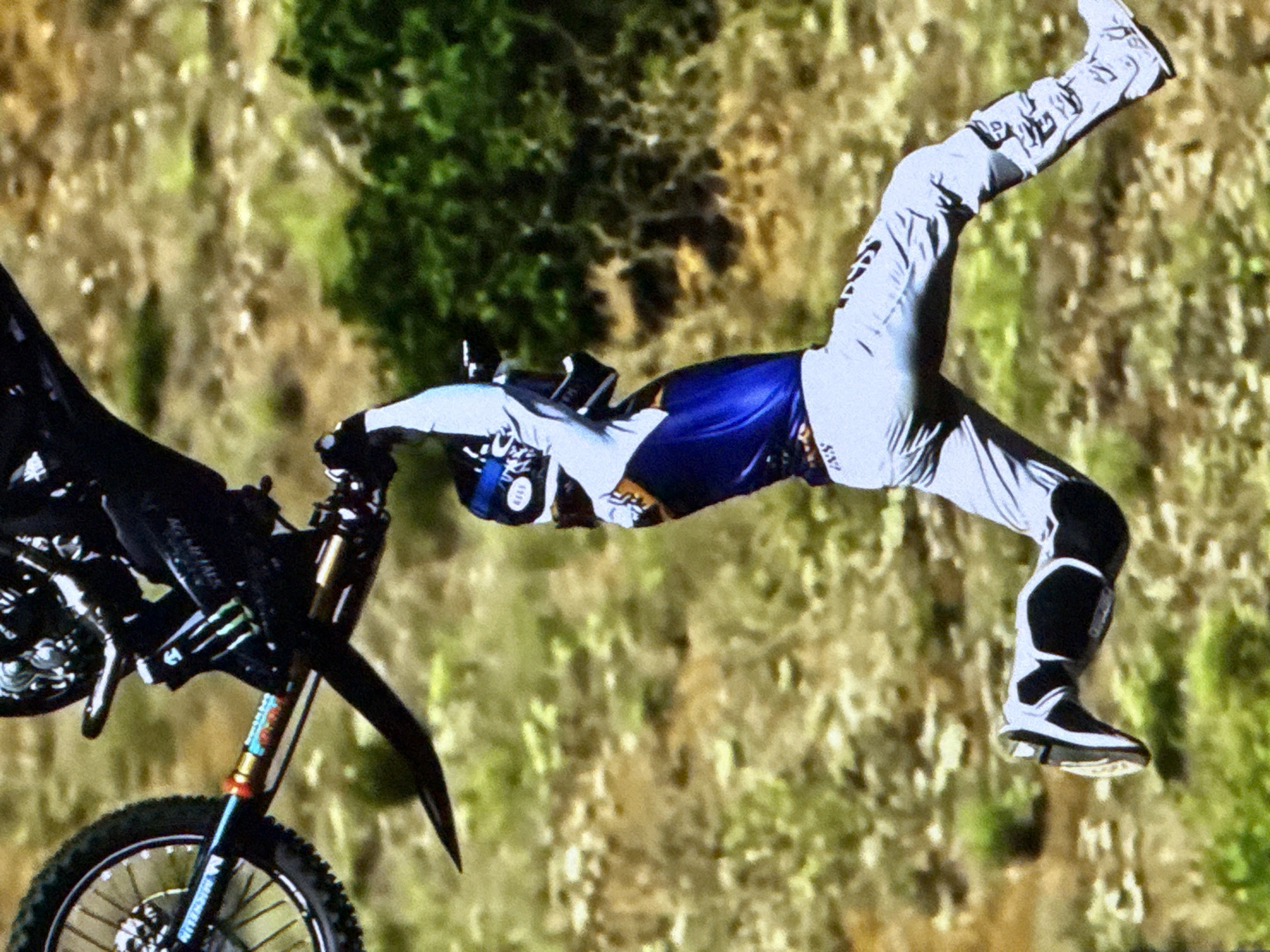 The image shows a motocross rider performing a stunt mid-air, with their body extended and one leg kicked out. The rider is wearing a blue and white jersey, grey pants, and white boots. The motorcycle is partially visible, with the front wheel and part of the frame in the upper left corner. The background is a blurred landscape of green vegetation and brown earth. The image appears to be a photograph of an action sport event. There is no indication of the location being the city of Cahul, Moldova.