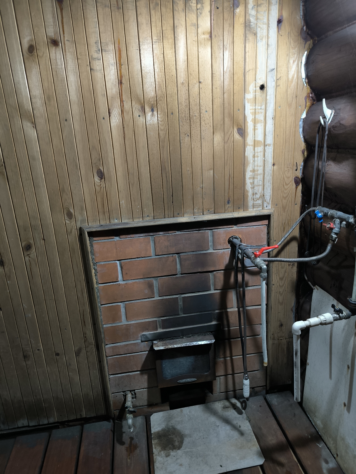 The image shows an interior scene, possibly a utility room or a part of a sauna, in a log-style building. The walls are primarily made of light-colored wooden planks, oriented vertically. In the center of the image, a recessed area is lined with red bricks, suggesting a fireplace or a stove surround. A metal ash door is visible within the brickwork. To the right, plumbing pipes with red and blue valves are installed, connected to flexible grey hoses. Some white plastic pipes are also present. The floor is made of dark wooden planks. The overall condition appears somewhat rustic and utilitarian. The location is specified as the city of Oktyabrsky, Russia.