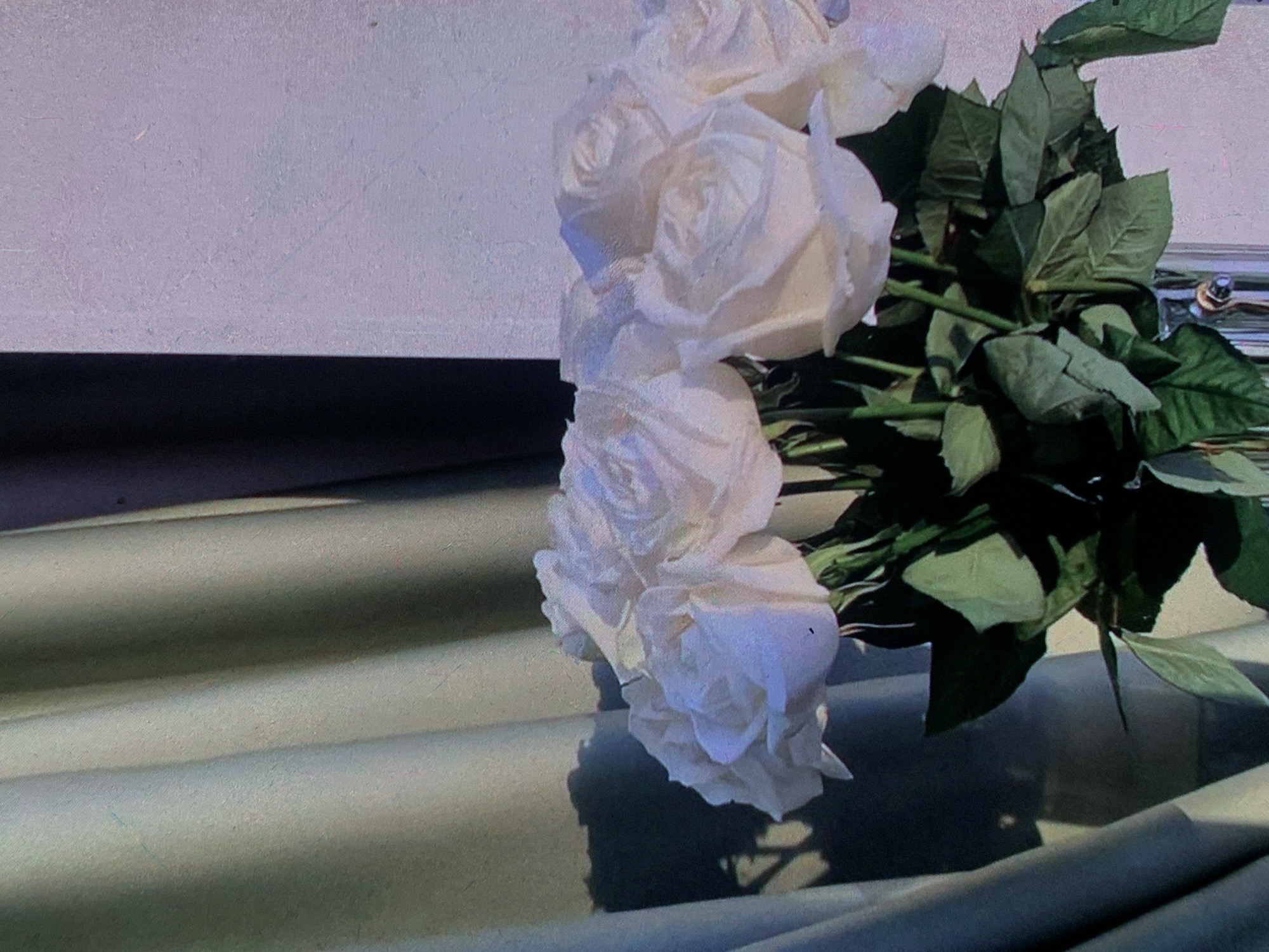 A bouquet of white roses is displayed in a clear glass vase. The roses are the primary subject, with their petals softly illuminated, creating a delicate and elegant appearance. Green leaves and stems are visible, adding a natural contrast to the white blooms.
The bouquet is positioned in front of a backdrop that consists of a light-colored curtain on the left and a textured wall on the right. The curtain is draped, creating folds and shadows that add depth to the scene. A dark shadow, possibly cast by an object or the roses themselves, falls on the curtain, creating an interesting visual element. The wall behind the roses appears to be a pale, possibly lavender or light gray, color with a subtle texture.
The overall lighting of the scene suggests it might be natural light filtering in from a window, possibly during the daytime. The quality of the image, with its slight graininess, could indicate it was taken with older film technology or a digital camera with a higher ISO setting.
There are no people or animals present in the image. The focus is entirely on the still life composition of the roses. The environment appears to be indoors, in a setting that could be a home, a floral shop, or an event space. There is no information within the image itself that would allow for a specific location in the city of Nizhny Tagil, Russia, to be identified.