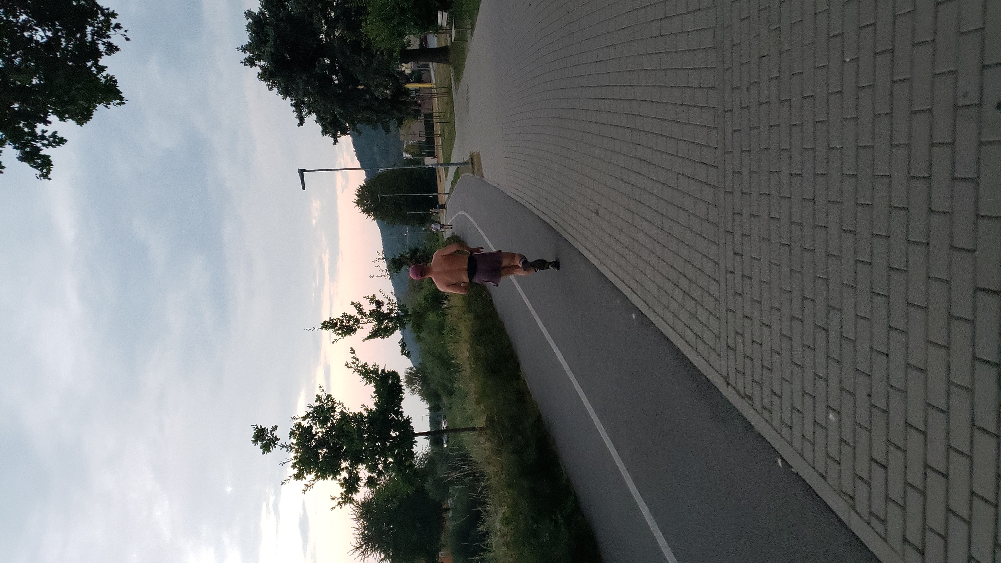 The image depicts a man running on a paved path next to a road with a white line. The man is shirtless, wearing purple shorts, a red cap, and running shoes. He is seen from behind, running away from the camera. The path is lined with trees and vegetation on the left, and a paved walkway with interlocking stones on the right. In the background, there are more trees, streetlights, buildings, and a hill. The sky is overcast with hints of pink and orange, suggesting it is either sunrise or sunset. The scene appears to be a recreational area in the city of Prešov, Slovakia, likely a park or a riverside path. There are a few other people visible in the distance on the path, also walking or running.