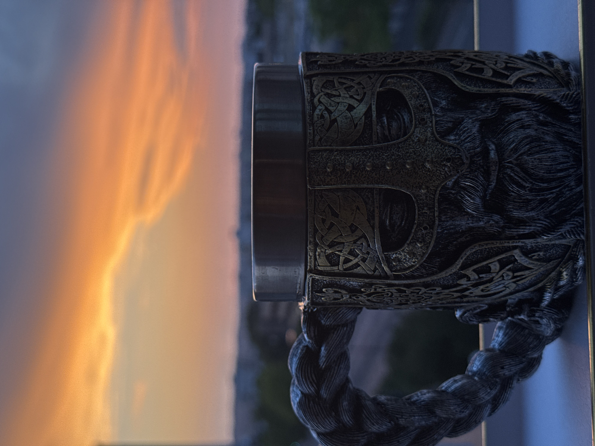 A close-up, eye-level, vertical shot captures a decorative mug with a Viking-themed design. The mug, made of a dark, metallic material, features intricate carvings of a Viking helmet and beard. The handle of the mug is designed to resemble a braided rope. It is placed on a flat surface, possibly a windowsill, with the top rim of the mug at the upper third of the frame.
In the background, a blurry cityscape unfolds under a dramatic sunset sky. The sky is dominated by vibrant oranges and yellows, with streaks of purple and blue indicating the fading light. The city lights are visible as blurred dots in the distance, and the silhouette of buildings and trees can be discerned against the colorful sky. The overall mood is serene and evocative, with the mug acting as a focal point against the expansive natural backdrop. The image is sharp and in focus on the mug, with the background artfully blurred to create depth.