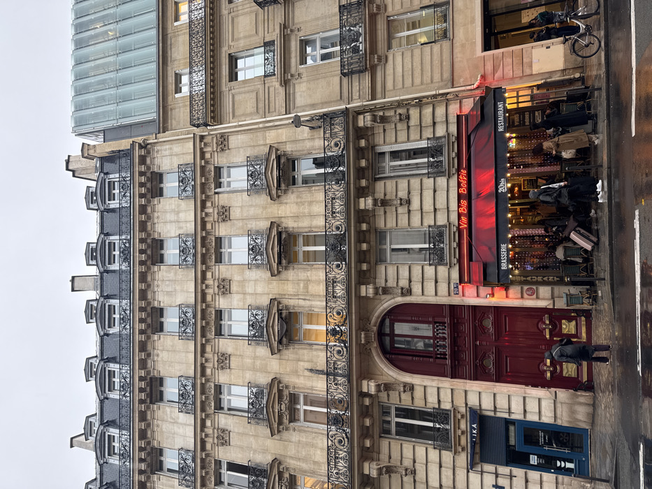 The image displays a street scene at 20 Rue La Boétie in Paris, France, under an overcast sky. The ground and street surfaces are wet, indicating recent precipitation.
The central feature is a multi-story building constructed of light-colored stone, characteristic of Haussmannian architecture. It possesses ornate wrought-iron balconies on the upper five visible floors, decorative cornices, and a dark grey mansard roof with several dormer windows and chimneys.
At street level, the building houses three distinct sections:
1. On the far left, a blue storefront with a sign reading "LIKA" is visible.
2. In the center, a large, dark red arched double-door entrance with brass kick plates and hardware stands prominently, featuring a small Juliet balcony above the doors.
3. To the right, the establishment named "Vin Bis Boétie" is identified by a red illuminated neon sign above a matching red awning. Additional signs read "BRASSERIE," "20bis," and "RESTAURANT." The interior is brightly lit with warm lights and string lights, revealing multiple patrons seated at tables.
On the wet sidewalk, several individuals are present. One person, wearing a dark coat, walks directly towards the dark red arched entrance. Under the awning of "Vin Bis Boétie," multiple individuals are standing or seated, some appearing to be holding or resting luggage, including a pink wheeled suitcase.
To the right of the central building, a contiguous structure of similar stone construction features balconies on its lower visible floors, while its upper section transitions into a contemporary, light-colored glass-paneled facade. A bicycle is parked on the sidewalk near this building, and another person is visible standing close by. Reflections from the building lights are visible on the wet street.