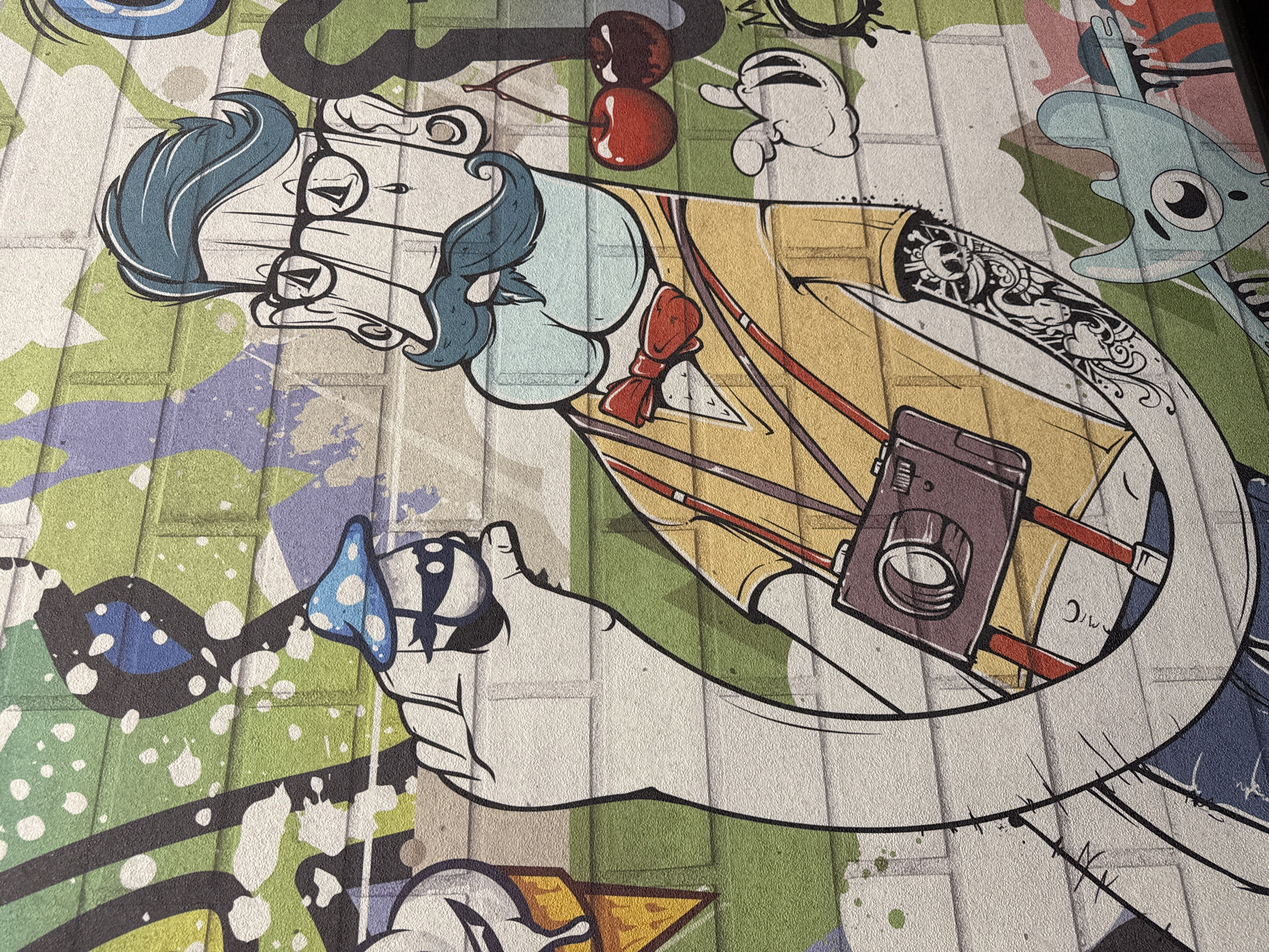 This media file captures a vibrant and detailed piece of urban art, likely a mural or graffiti, displayed on a textured surface resembling a block or brick wall. The artwork features a stylized, cartoon-like male figure as its central subject. He has blue hair, a distinctive beard, and wears round, thick-rimmed glasses, a light blue shirt, a yellow jacket, and a red bow tie.
The figure is depicted holding a vintage-style camera with a strap in his left hand. His right hand holds a small, blue object adorned with white polka dots, possibly a mushroom or a bell. His left arm (on the right side of the image) is covered in an intricate sleeve tattoo featuring skulls, organic patterns, and what appear to be creatures like dragons or serpents.
Around the main character, several other cartoon elements are visible: a pair of bright red cherries with stems in the upper middle, a small white ghost-like creature with hands, and a light blue, amorphous blob-like character with a single eye to the right. The background consists of abstract splashes and blocks of color, predominantly green, purple, and blue, accented with white splatter effects, creating a dynamic and energetic backdrop. The rough, underlying texture of the wall is evident throughout the painting, giving it a tangible depth. The artistic style is bold and graphic, characterized by thick outlines and bright, contrasting colors. There is no visible text in the image. This artwork is located in Yekaterinburg, Russia.