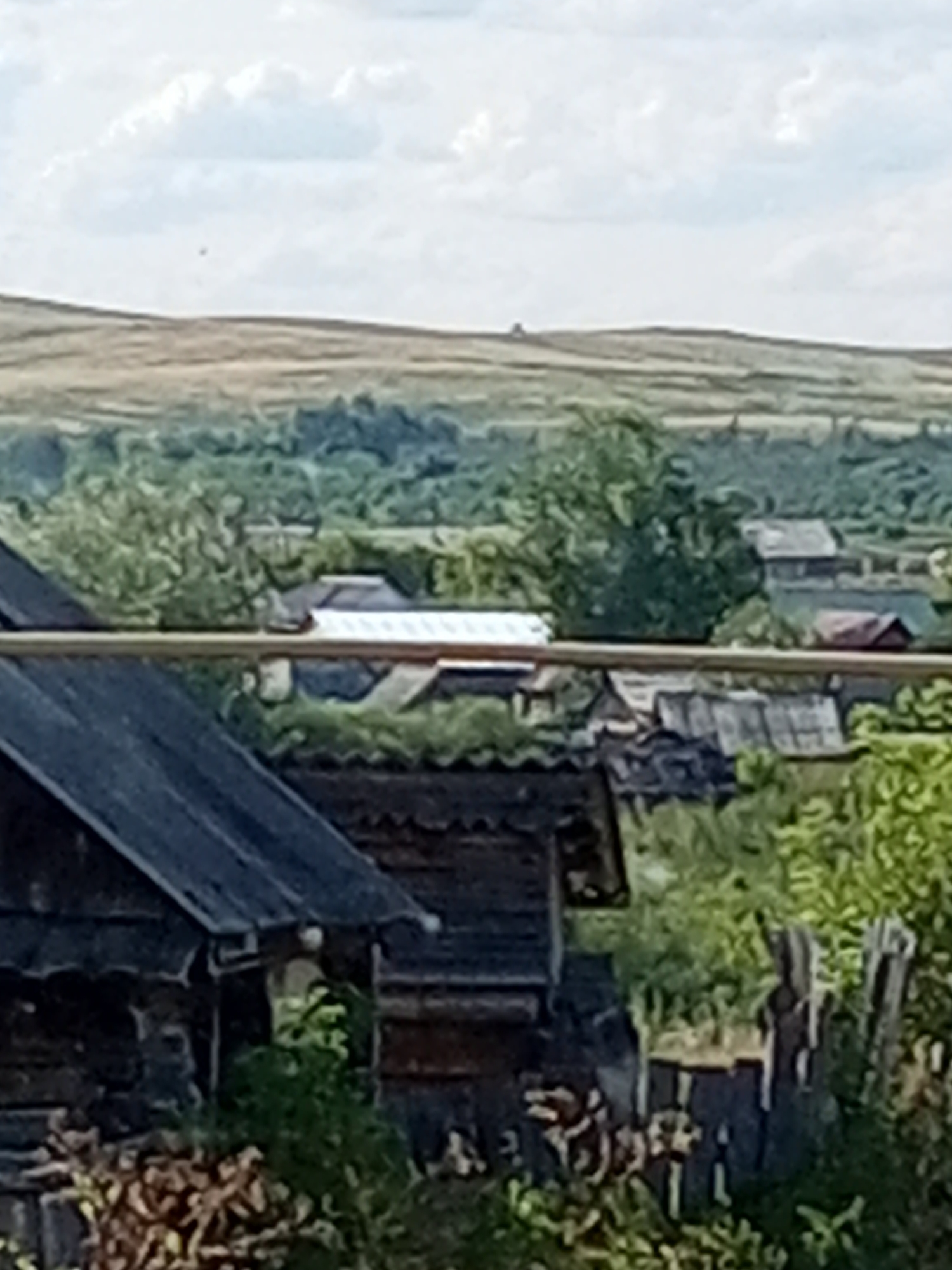 The image shows a rural landscape with several wooden structures and vegetation. In the foreground, on the left, is a portion of a dark-colored wooden building with a slanted roof. To the right of this building, there is a stack of chopped wood. Further back, a horizontal wooden beam runs across the frame. Behind the beam, a cluster of wooden structures with various roofs are visible, some appearing to be sheds or small houses. Green trees and bushes are interspersed among these buildings, and beyond them, a gently sloping, open field extends towards a distant, hazy horizon under a cloudy sky. The scene is situated in the village of Kuyantaevo, Russia. There are no visible people in the image.
